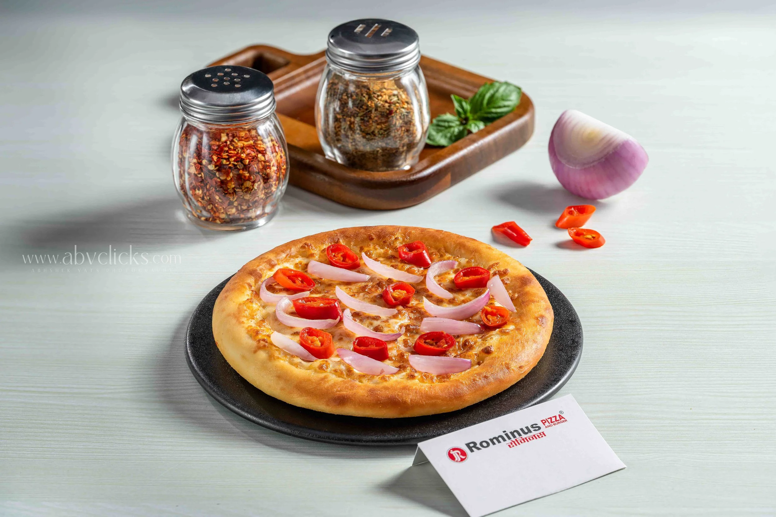 A pizza with sliced red chili peppers and onions on a black plate, placed on a light-colored surface with a table card that has the logo of Rominu's pizza restaurant.