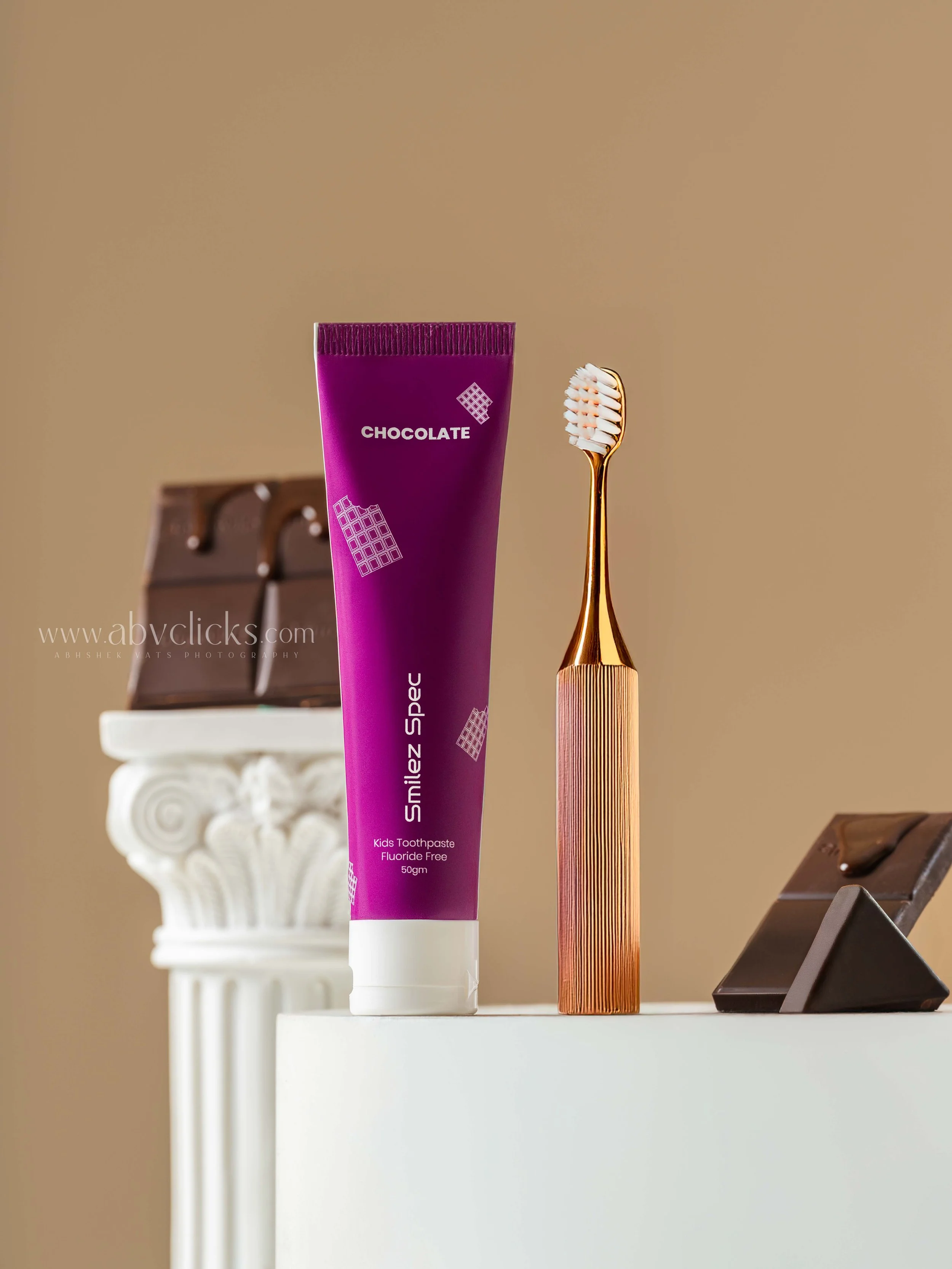 A tube of kids fluoride-free toothpaste labeled 'Smilez Space' in purple, a gold toothbrush with white bristles, and chocolate bars on a white surface with beige background.