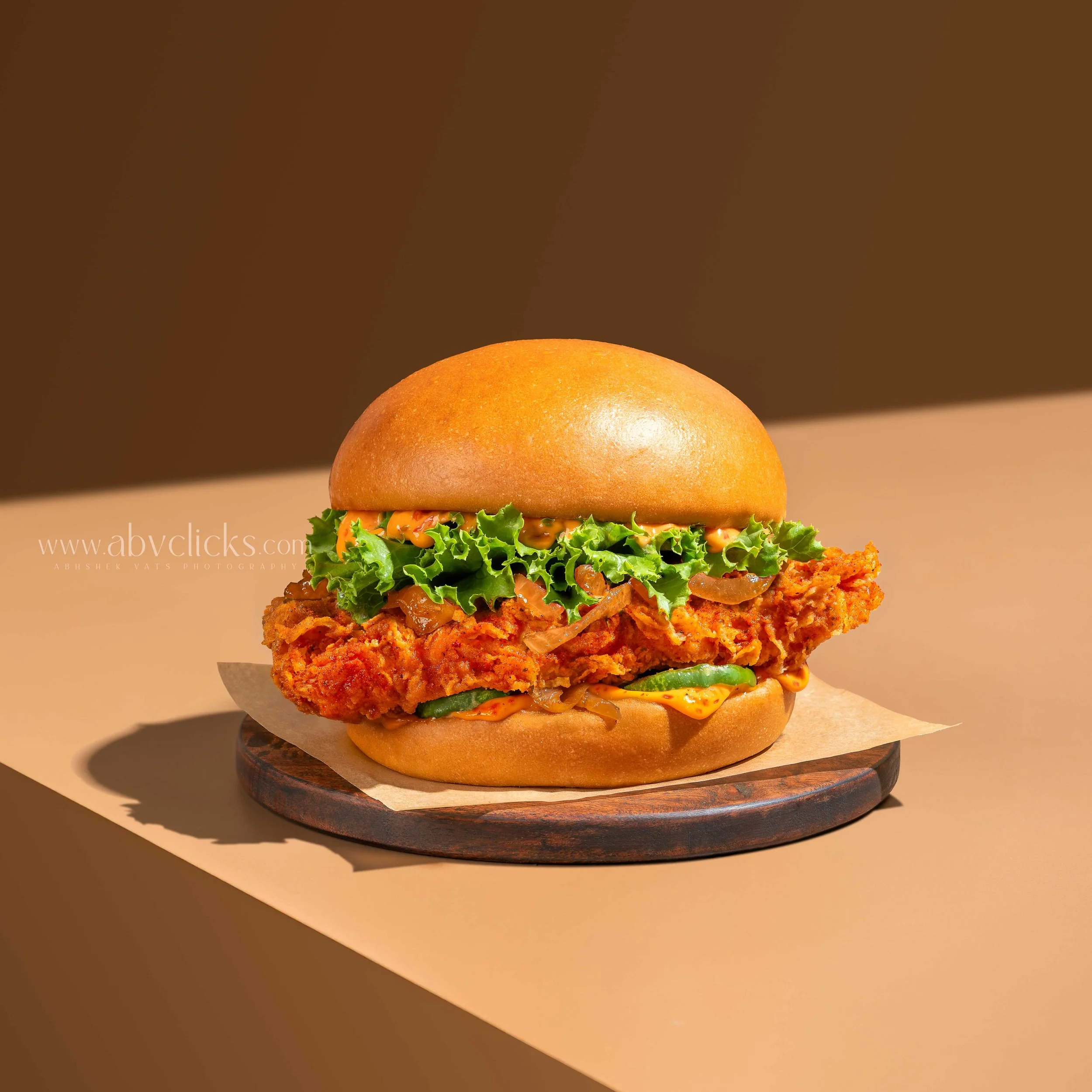 For brand Sandburgs, Close-up of a burger with crispy fried chicken, lettuce, and sauce in a bun, placed on a wooden round board with a light brown background.