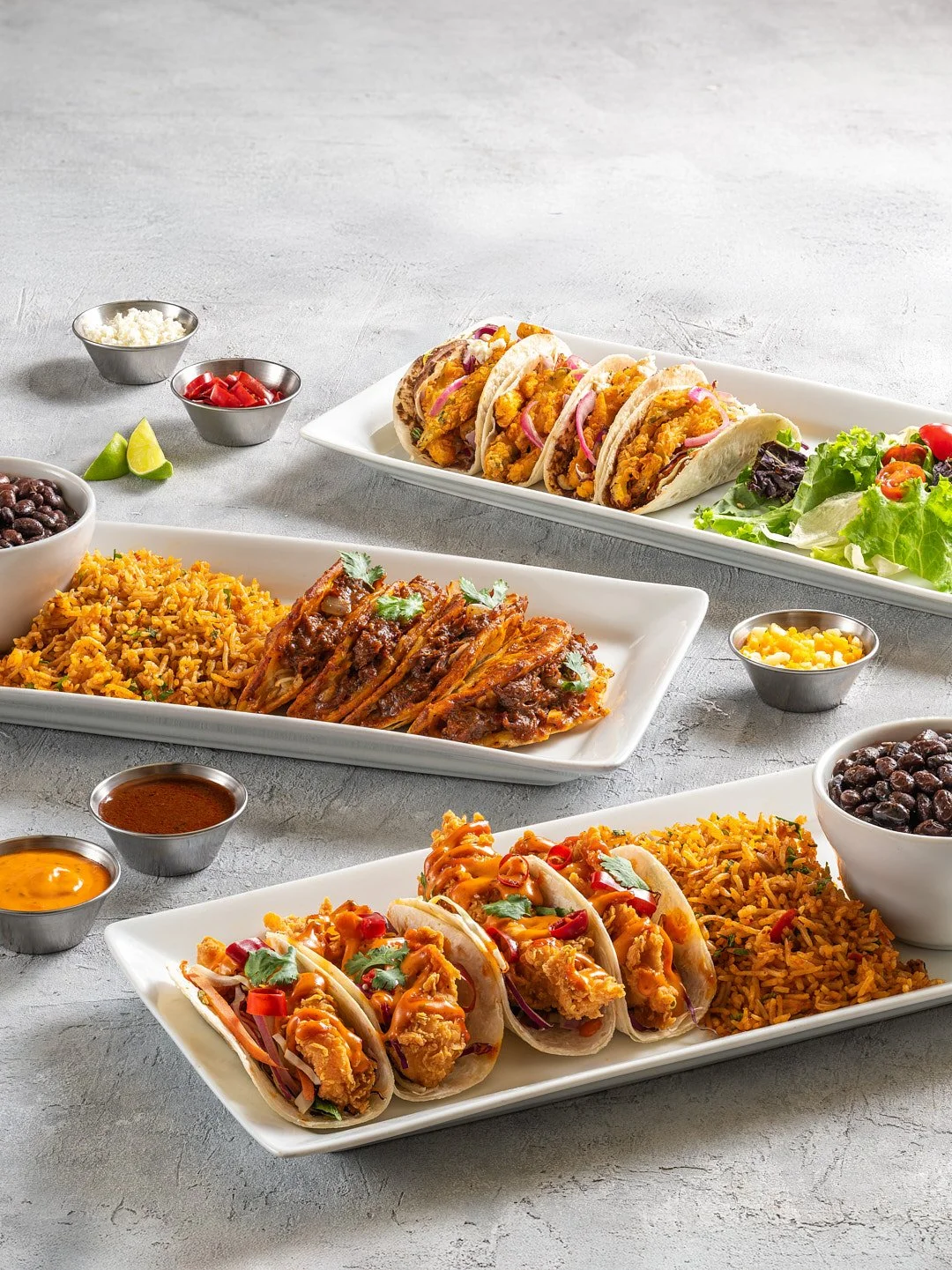 Three white rectangular plates with tacos, rice, and grilled meat, garnished with cilantro, served with small bowls of salsa, beans, and sauces, and a side salad with lettuce and cherry tomatoes, on a gray textured surface.
