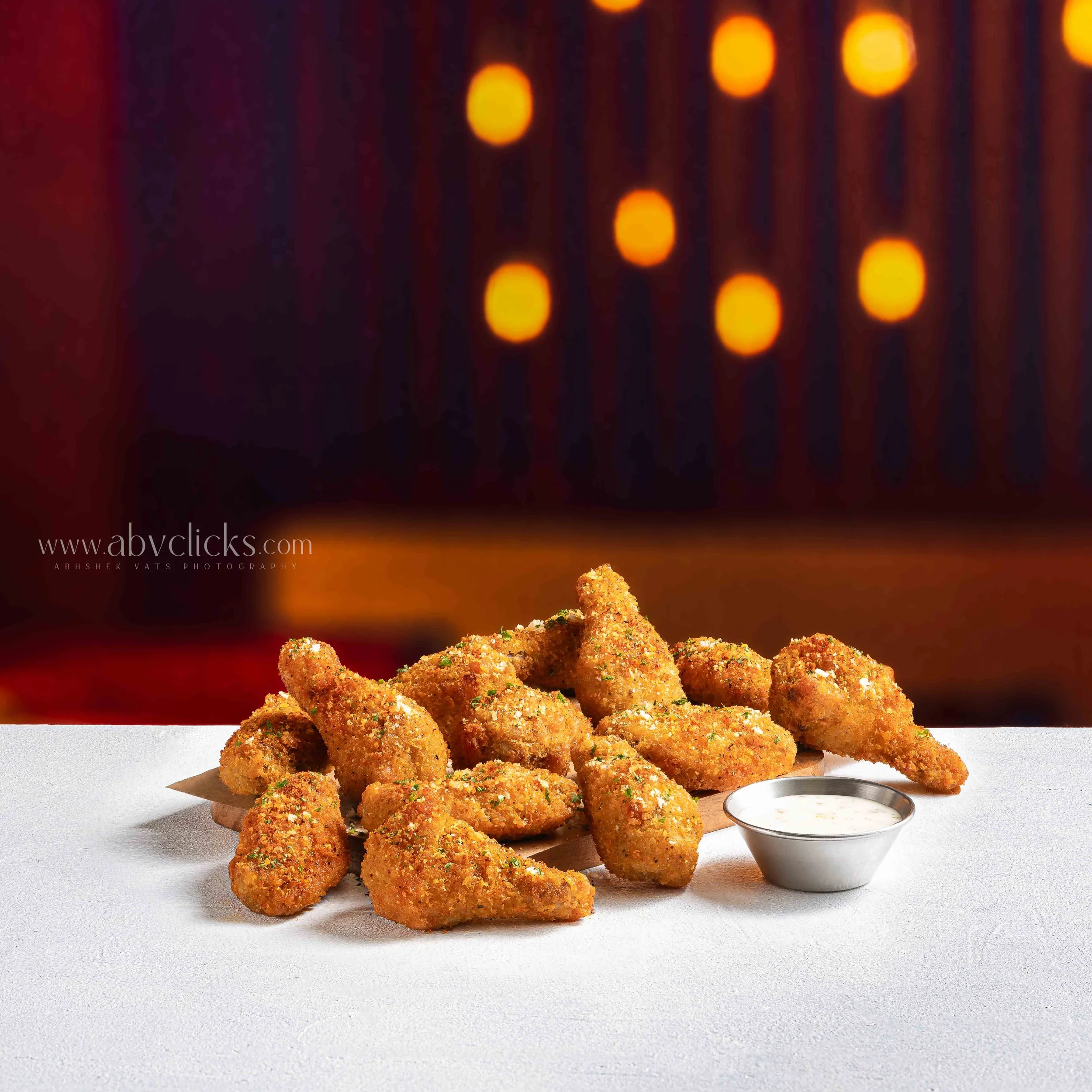 Crispy breaded chicken nuggets on a white surface with a small bowl of dipping sauce, against a blurred dark background with orange lights.