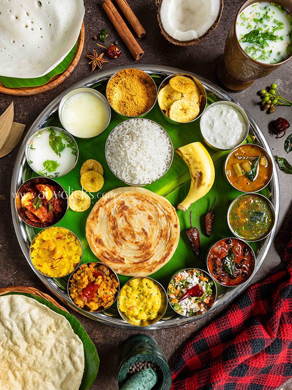 Traditional South Indian feast on a large metal tray with banana leaf lining, featuring rice, chapati, various lentil and vegetable curries, chutneys, pickles, bananas, and buttermilk, surrounded by additional naan, coconut, and spices.