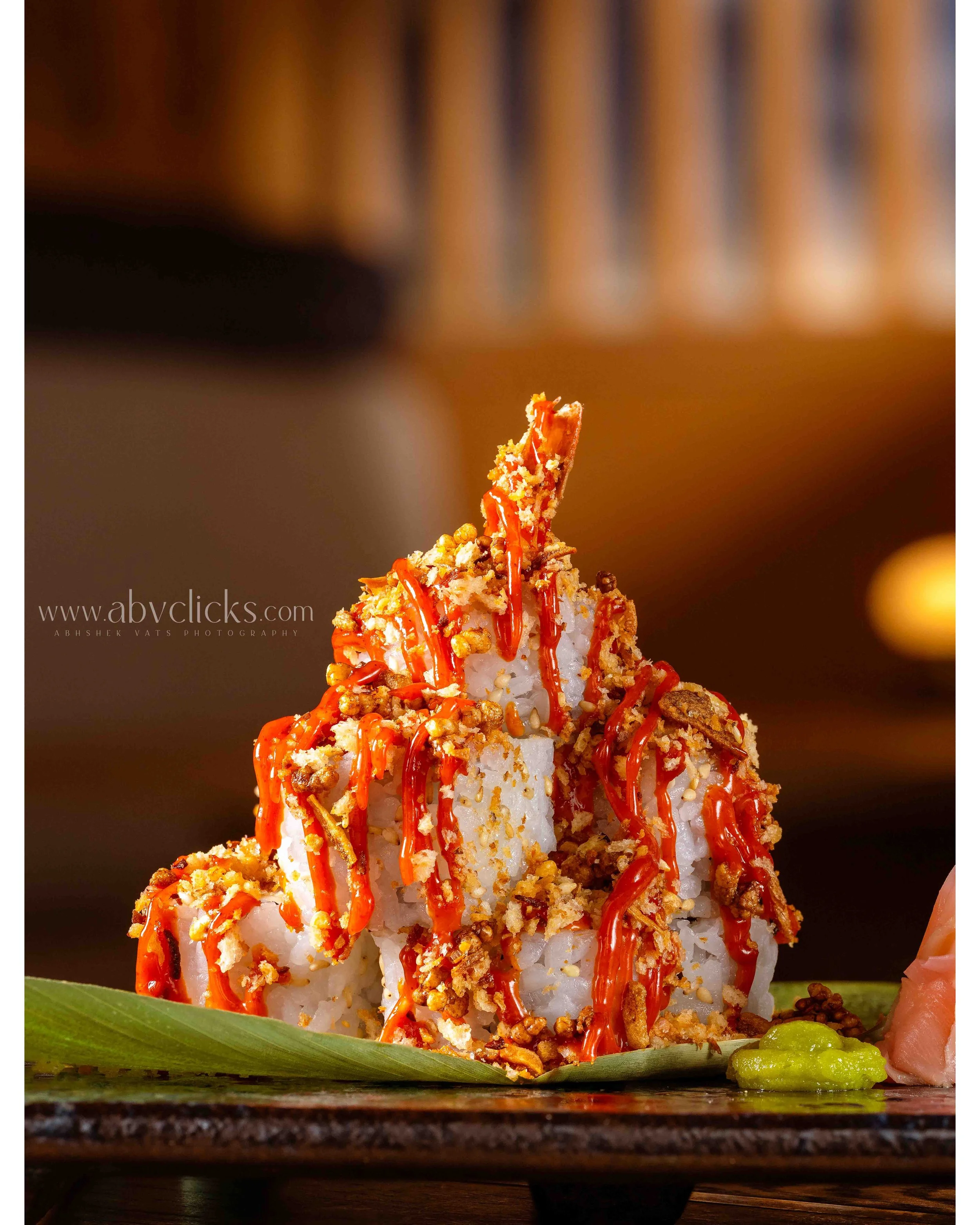 Close-up of a sushi roll topped with spicy sauce and crunchy toppings, with a green leaf and wasabi on the side.