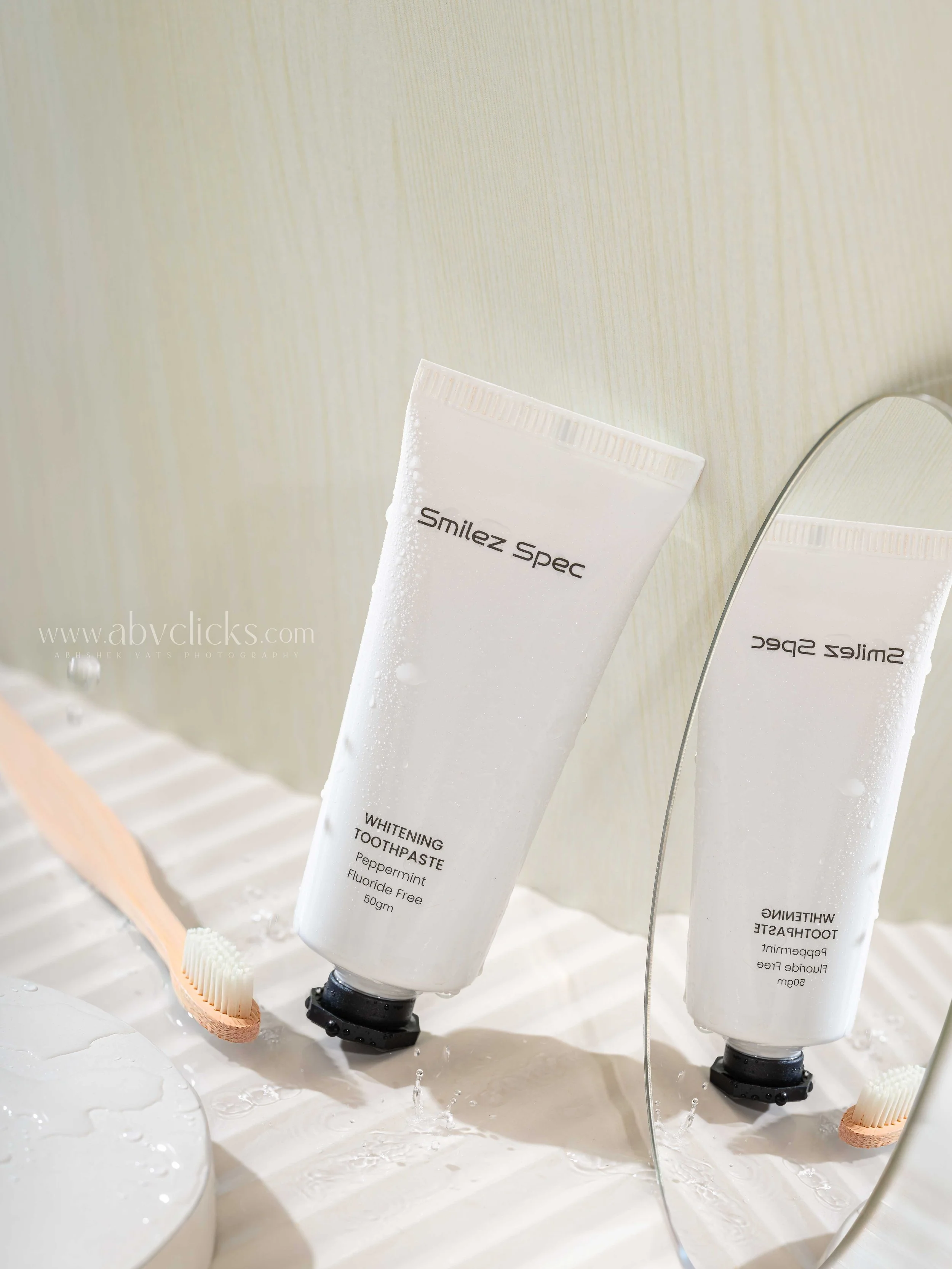White tube of Whitening Toothpaste labeled 'Smilez Spec' with a toothbrush leaning against it, in a bathroom setting with water droplets and reflection in a mirror.