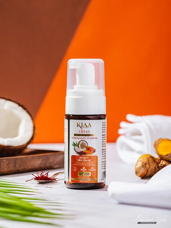 A brown skin-colored bottle of KIAA UBTAN face wash with turmeric and saffron, placed on a white surface with coconuts, ginger roots, and white towels around it, and an orange background.