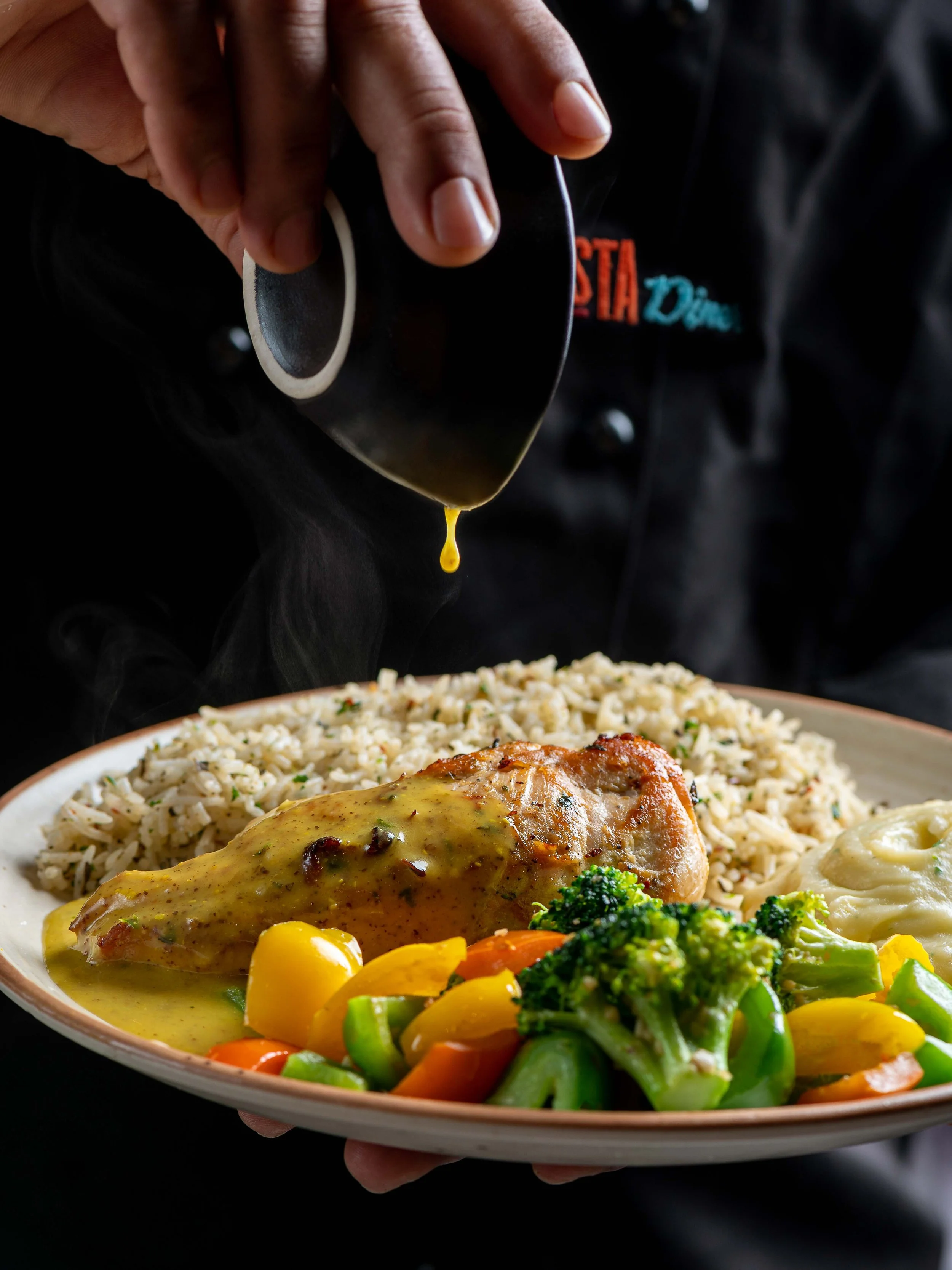 A person serving sauce over a plate of rice, chicken with gravy, vegetables, and mashed potatoes.