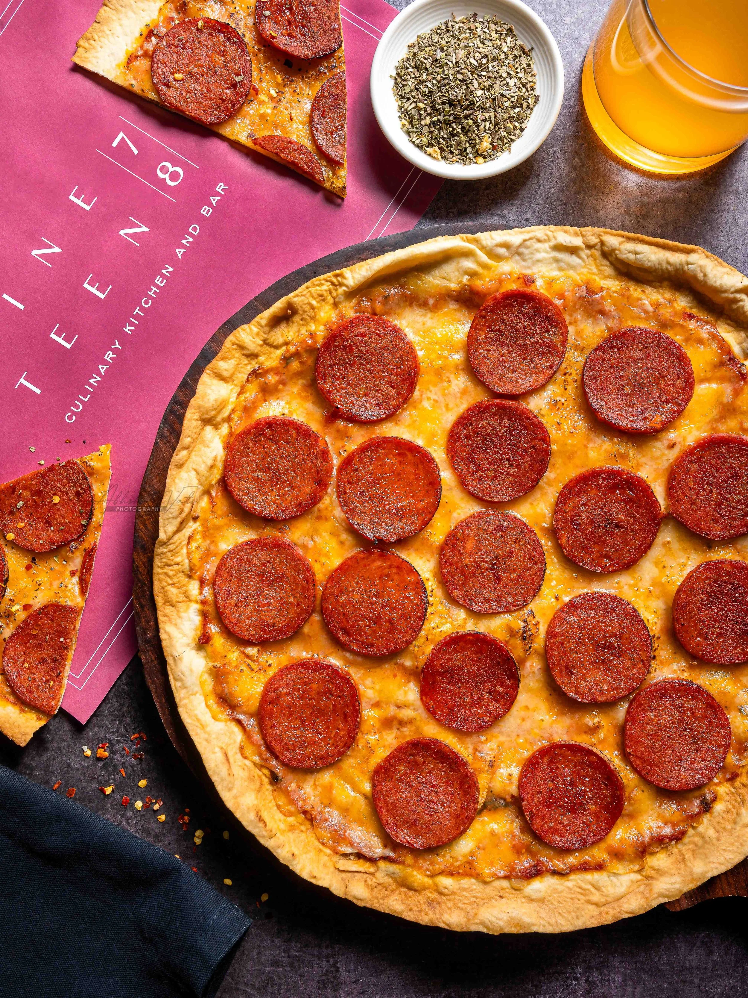 Close-up of a pepperoni pizza on a black tray, with a glass of beer, a bowl of chopped herbs, and a pepperoni slice on pink and gray background.