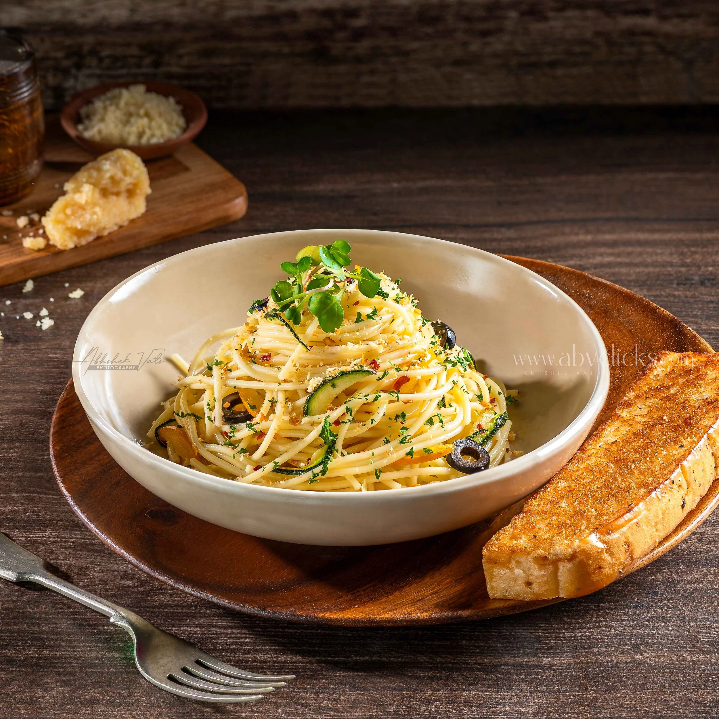 A bowl of pasta garnished with herbs and vegetables, placed on a wooden tray with toasted bread and salt and pepper shakers on a dark wooden table.