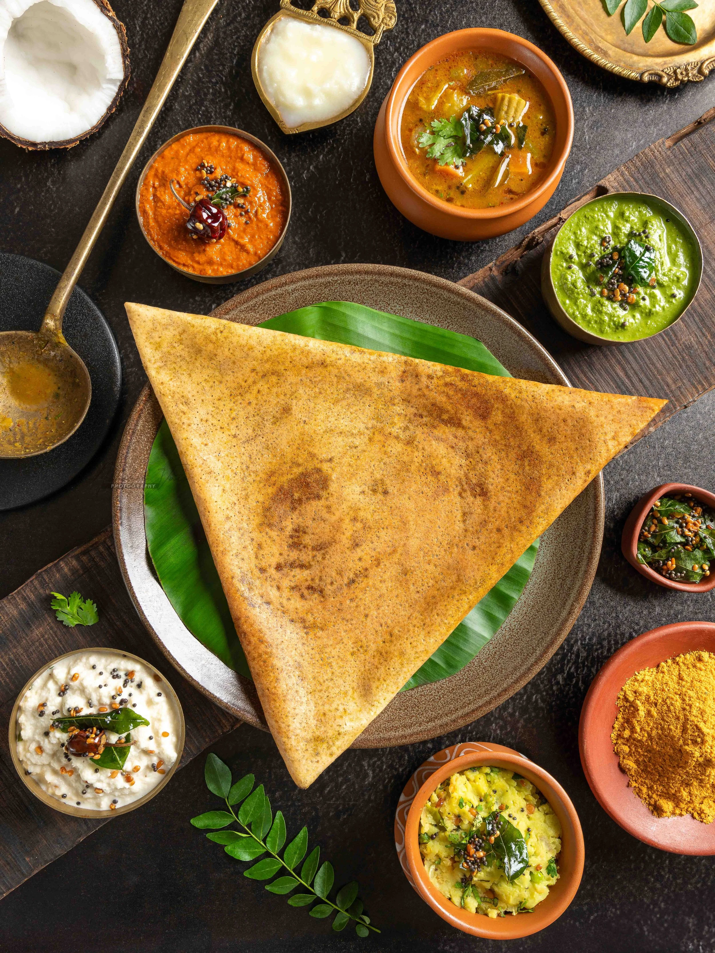 A traditional Indian meal featuring a triangular large dosa, bowls of sambar, coconut chutney, tomato chutney, and potato curry, garnished with cilantro and black mustard seeds.