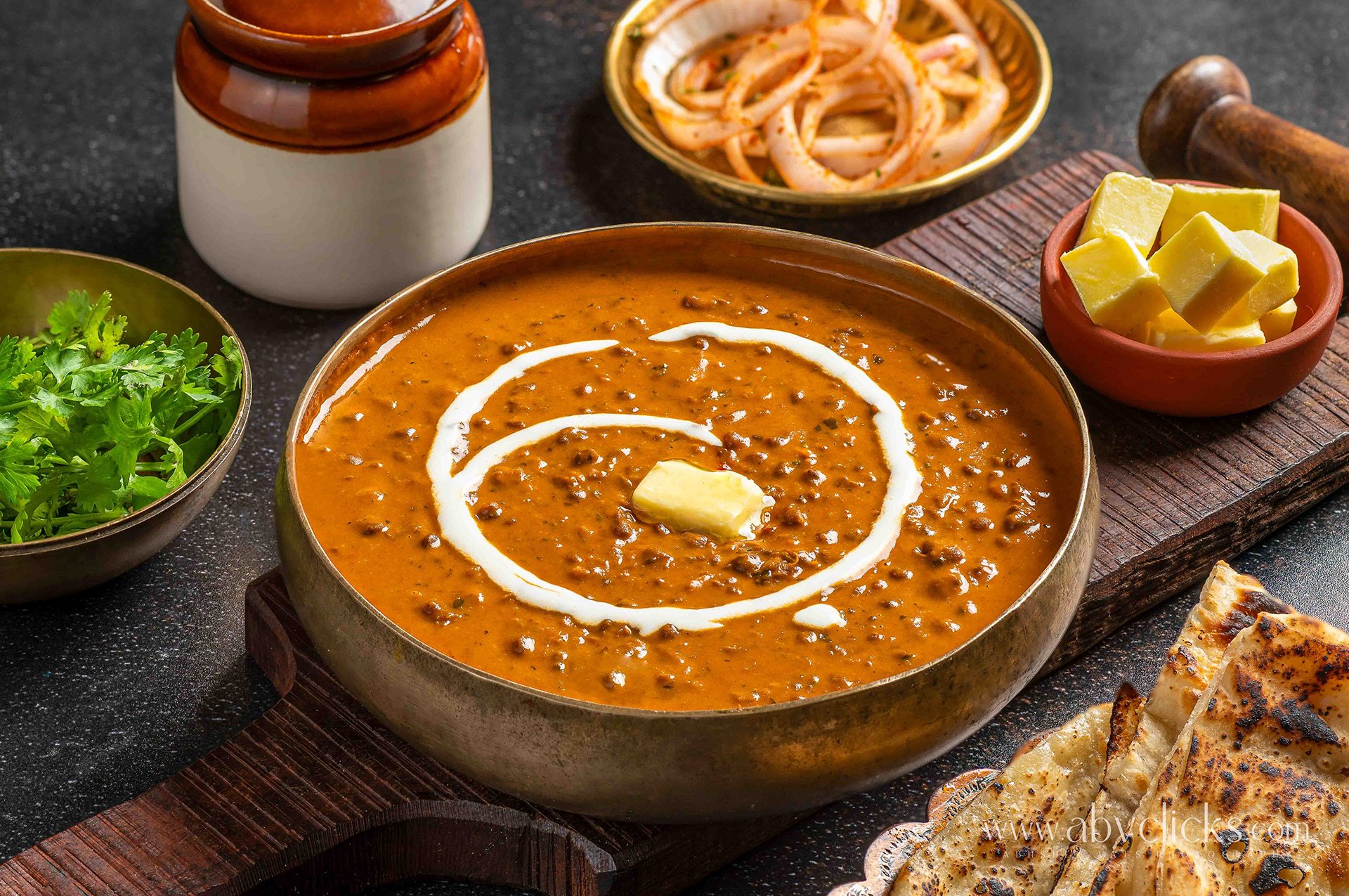 For brand Dana Choga gurgaon, a bowl of Indian lentil dal garnished with butter and cream, with side dishes including chopped green herbs, pickled onions, and chopped butter, served on a wooden tray with grilled bread and a rustic cutting board.