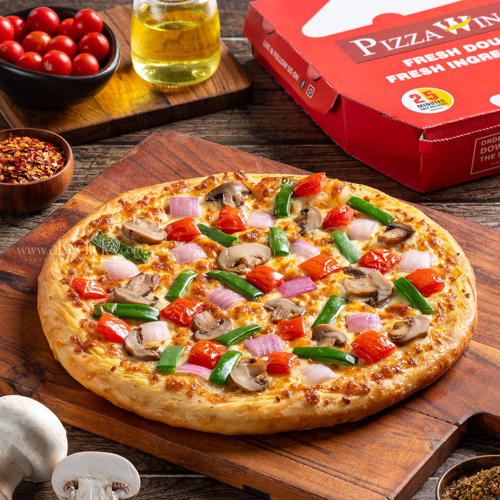 A pizza with cheese, green peppers, cherry tomatoes, mushrooms, and onions on a wooden board, with cherry tomatoes, olive oil, crushed red pepper, and garlic in the background.