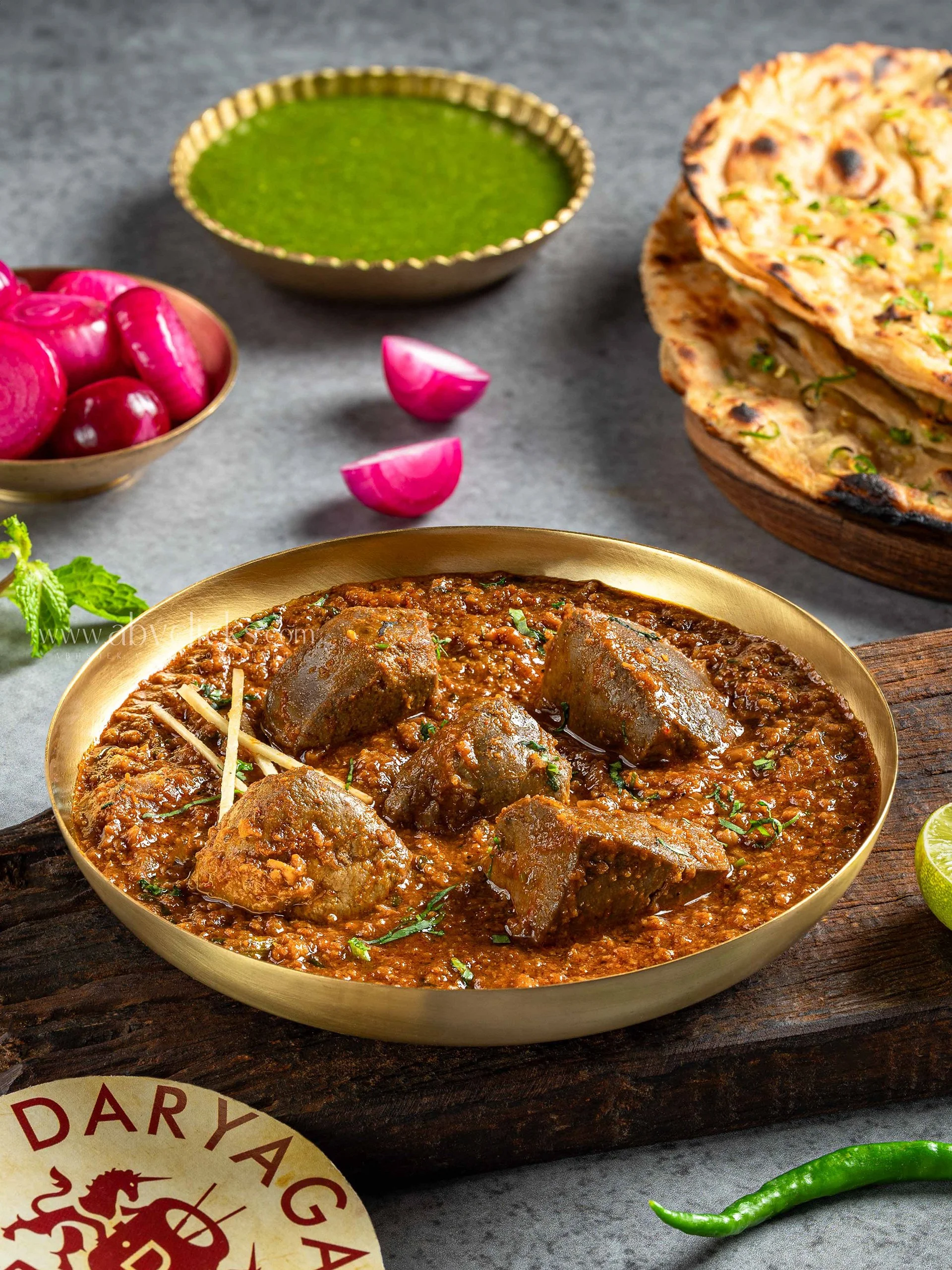 A bowl of Indian curry with chunks of meat and spices, accompanied by radish slices, green chili, and naan bread topped with herbs, on a rustic wooden surface.