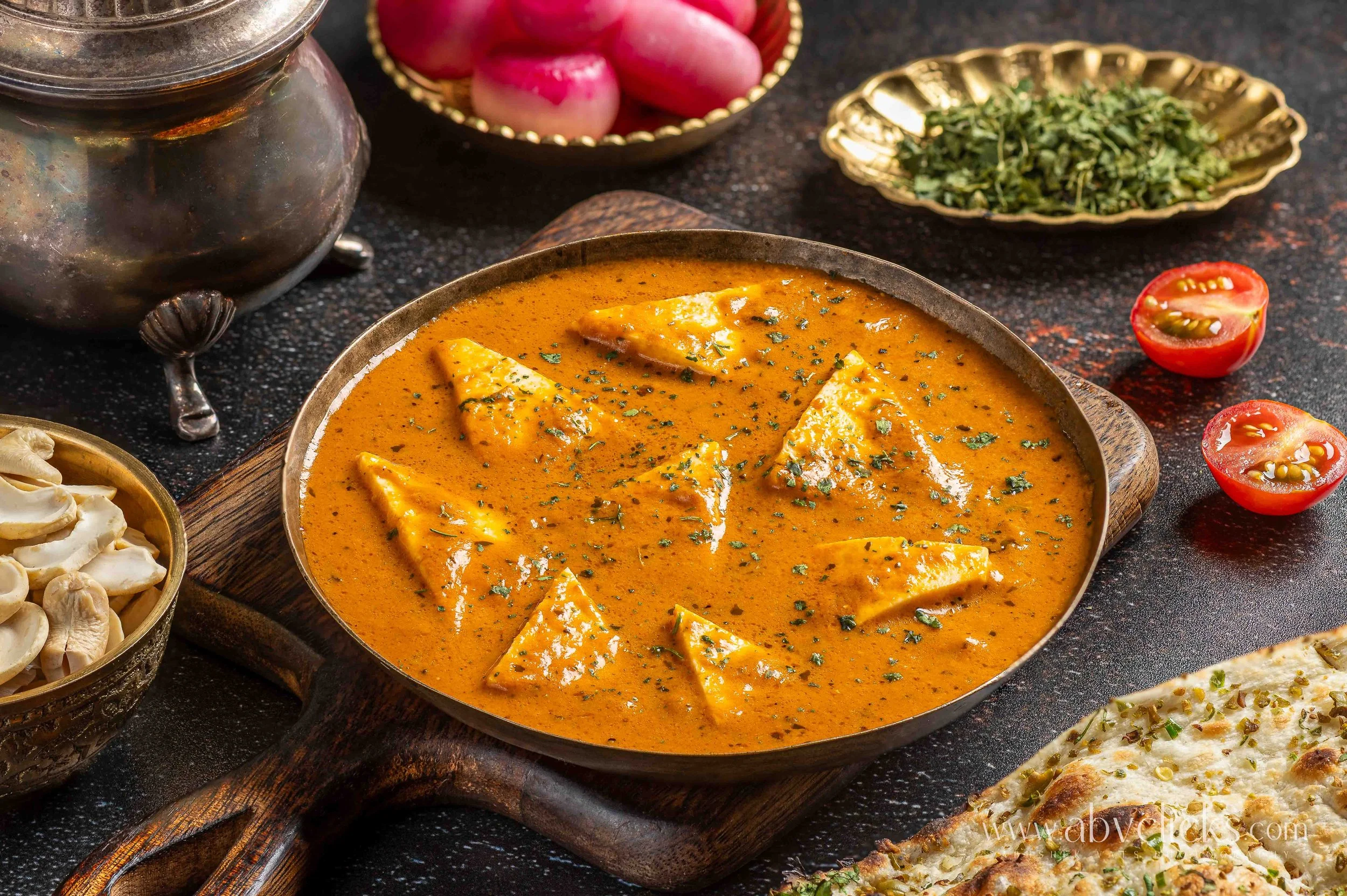 A bowl of Indian chicken curry garnished with herbs, surrounded by vegetables including cherry tomatoes, radishes, mushrooms, and flatbread with pistachios on top, all displayed on a dark surface.