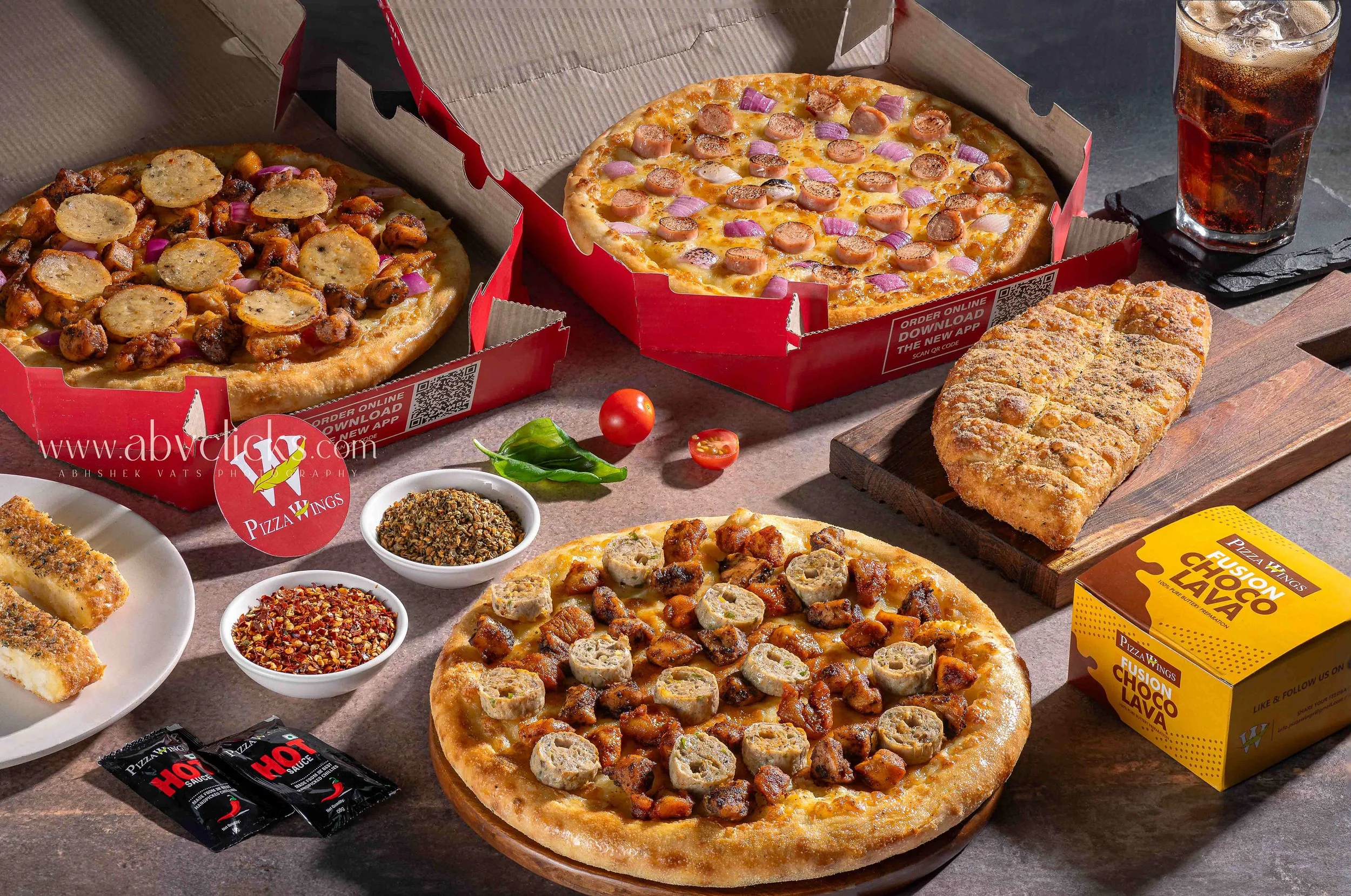 Three pizzas in open boxes on a table, with a glass of cola, spices, chopped tomatoes, and a box of chocolate-flavored chips nearby.