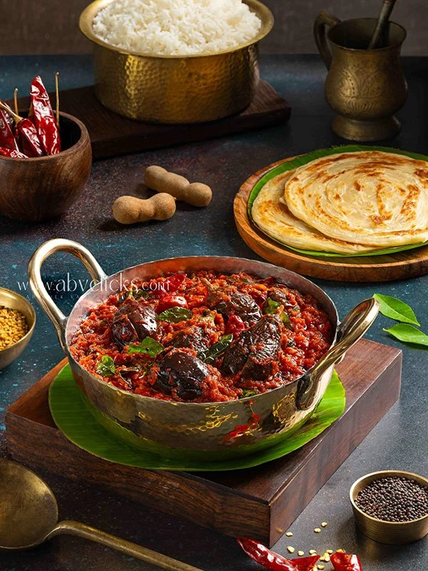 A traditional Indian meal with a bowl of white rice on a dark wooden board, a plate of crispy parathas, and a metal bowl of spicy vegetable curry surrounded by spices and condiments on a dark surface.