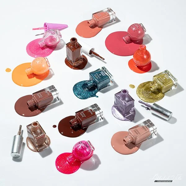 Multiple bottles of nail polish in various colors, some open with brushes, and some with spilled polish on a white surface.