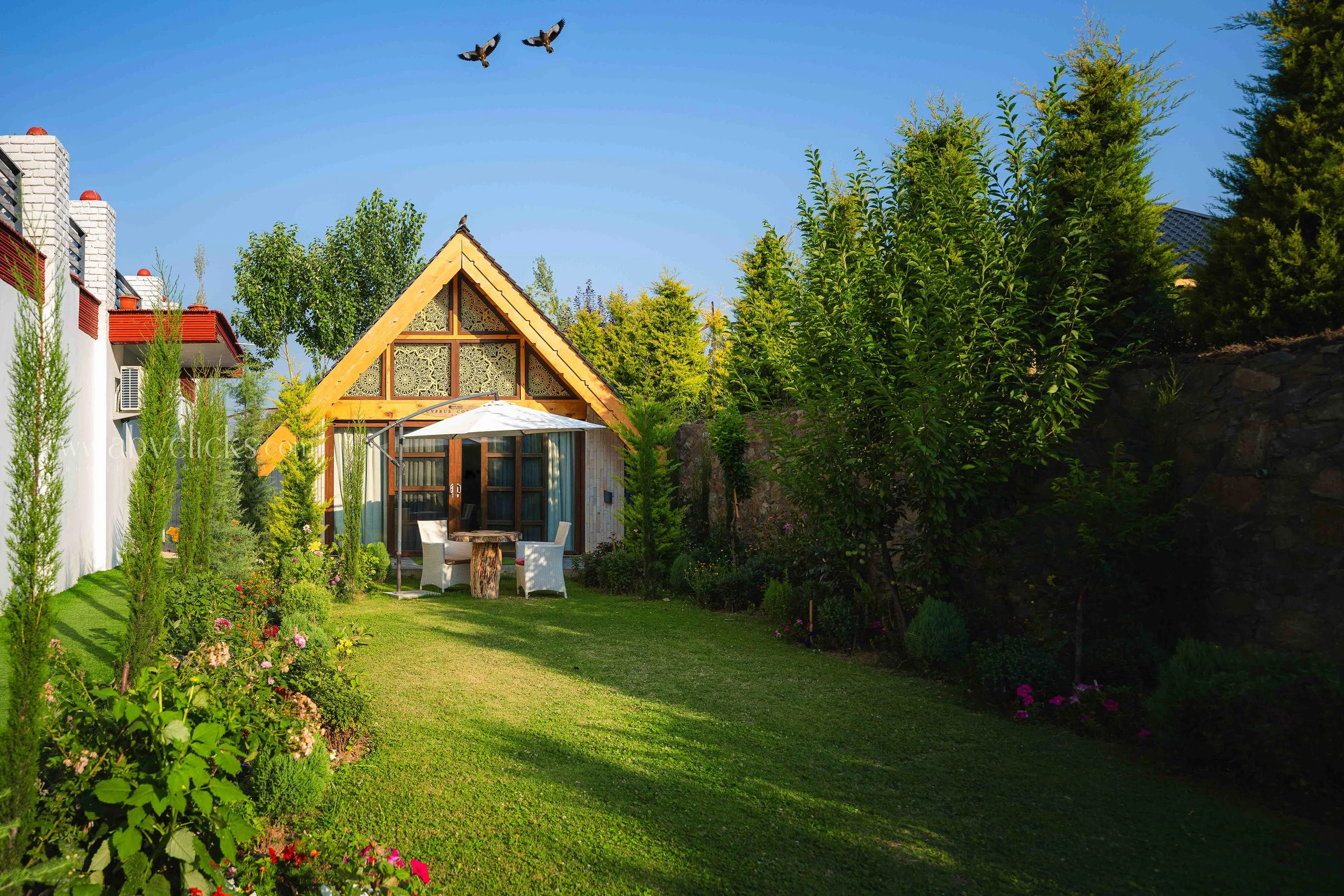 A beautiful backyard garden with lush green grass, blooming flowers, tall trees, a glass door patio area with outdoor dining furniture, and a wooden A-frame house in the background. Two birds are flying in the clear blue sky.