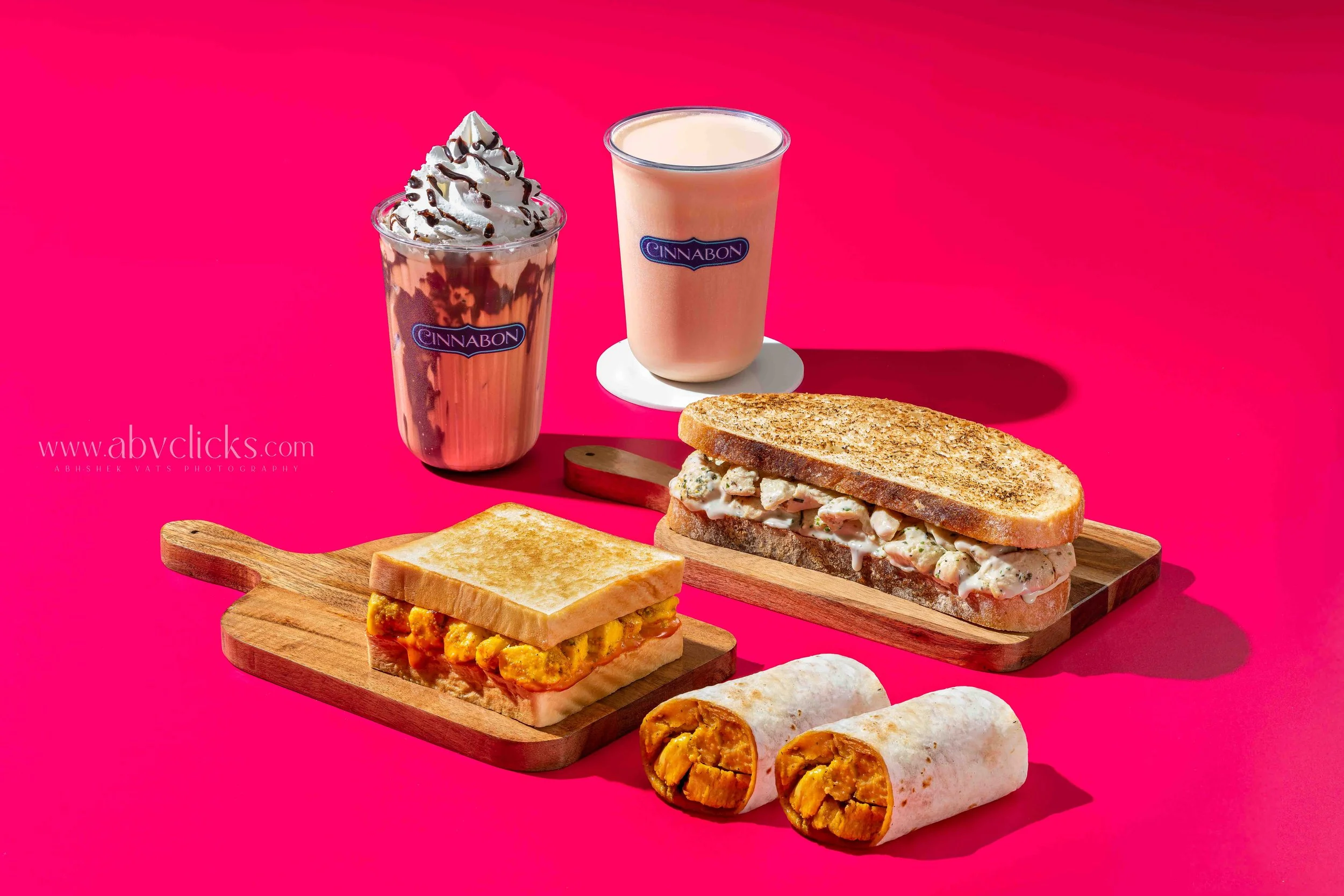 Two milkshakes, one topped with whipped cream and chocolate drizzle, and the other plain, along with a sandwich on toasted bread, a wrap, and a mini toasted sandwich on a wooden serving board, set against a pink background.