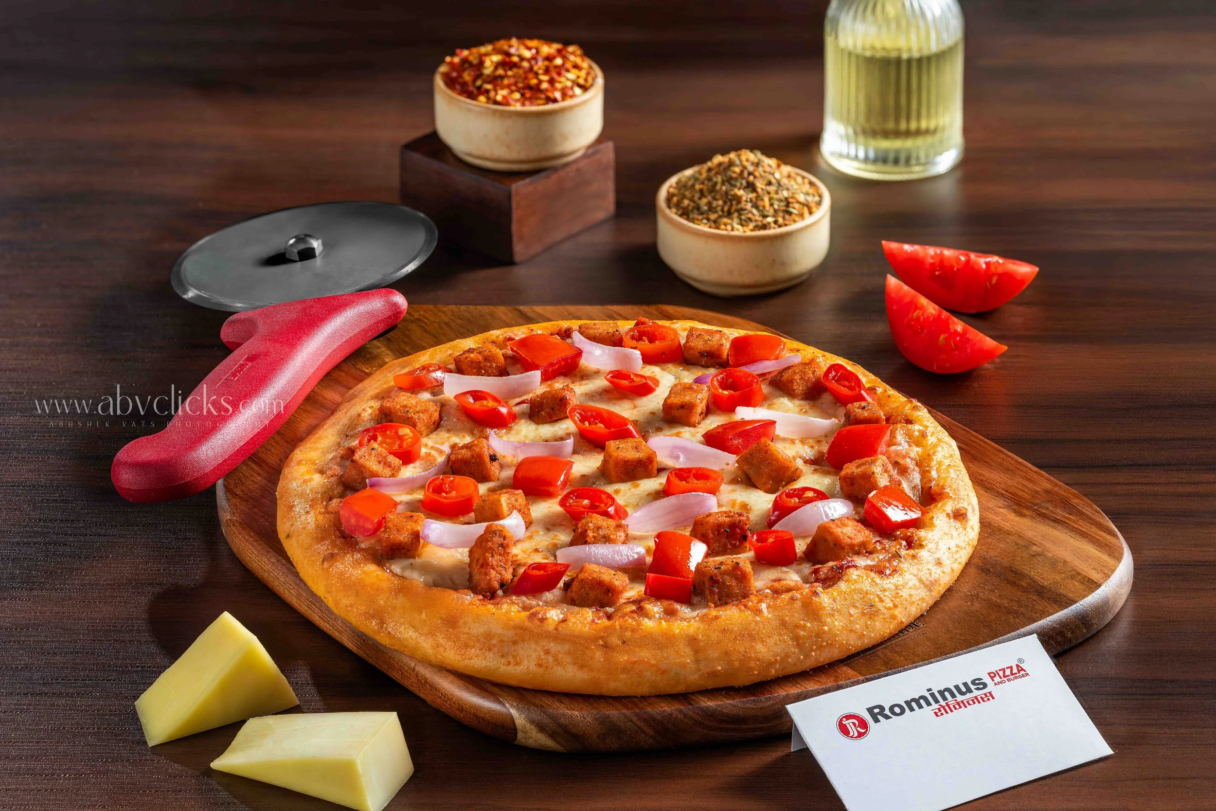 Close-up of a pepperoni and sausage pizza on a wooden board with a red pizza cutter and slices of cheese nearby. In the background, there are bowls of crushed red pepper flakes and herbs, a glass bottle of oil, and sliced tomatoes on a dark wooden ta
