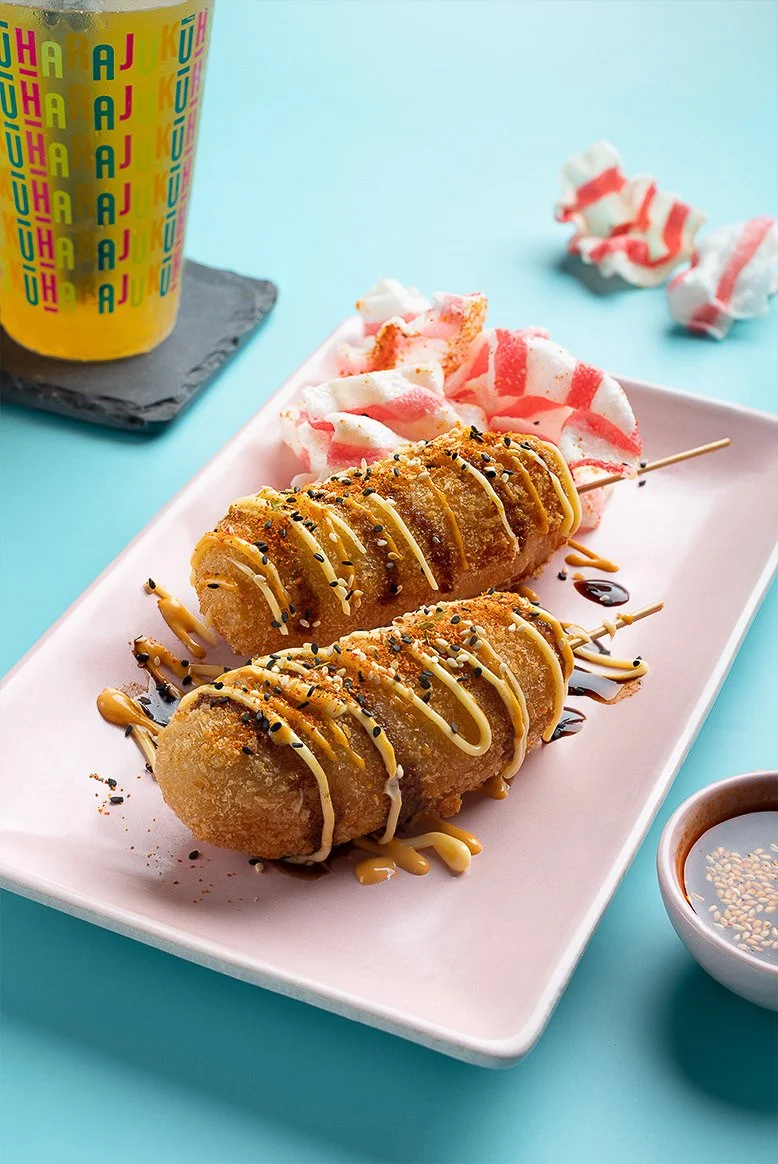 Two crispy spring rolls drizzled with sauces and sprinkled with sesame seeds serve on a pink rectangular plate, accompanied by pink and white striped marshmallow snacks and a colorful beverage in a glass cup with a straw, on a light blue table.