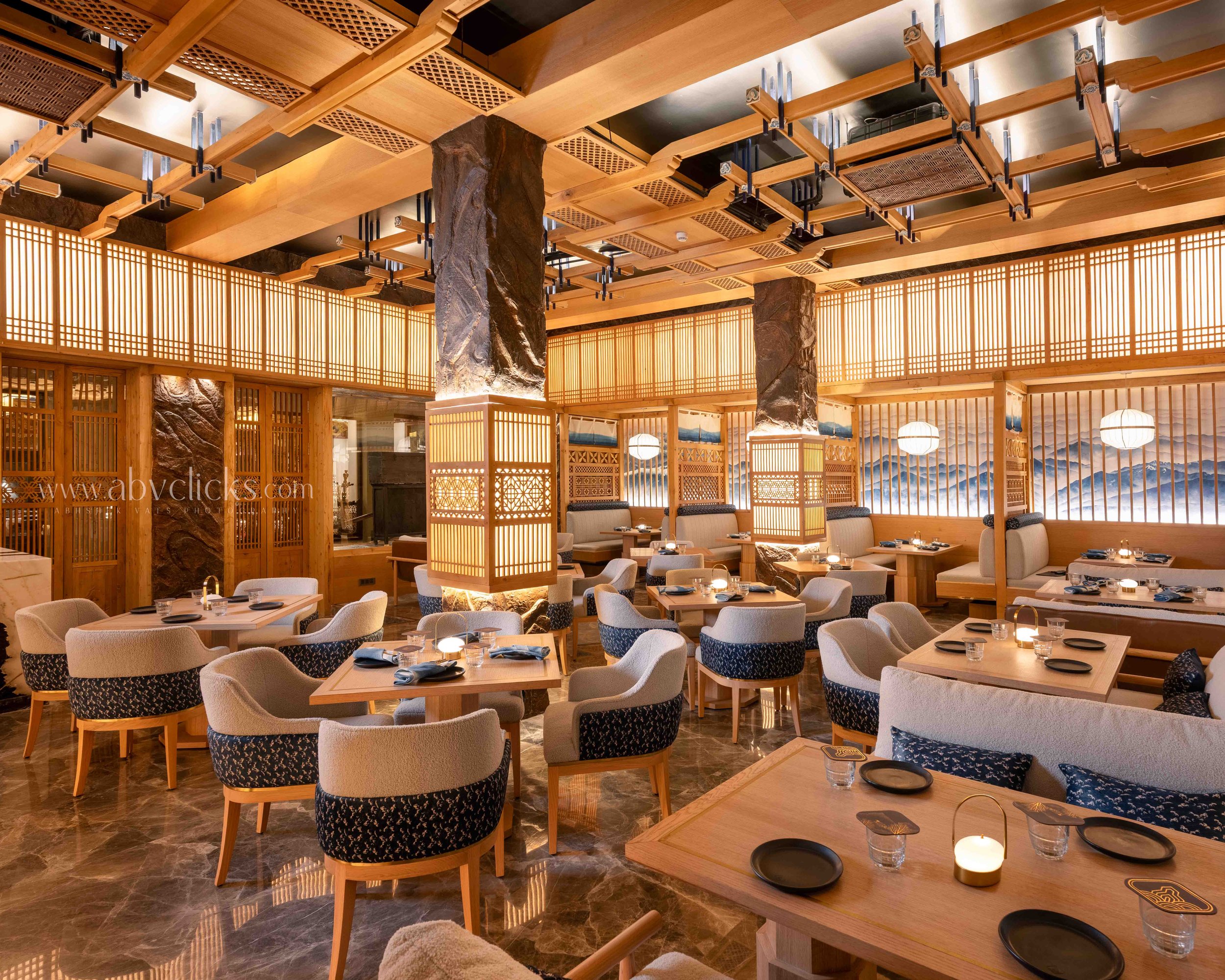 Interior of a restaurant with wooden decor, low tables set with black plates and glasses, plush upholstered chairs, and decorative lighting including lanterns and candles.