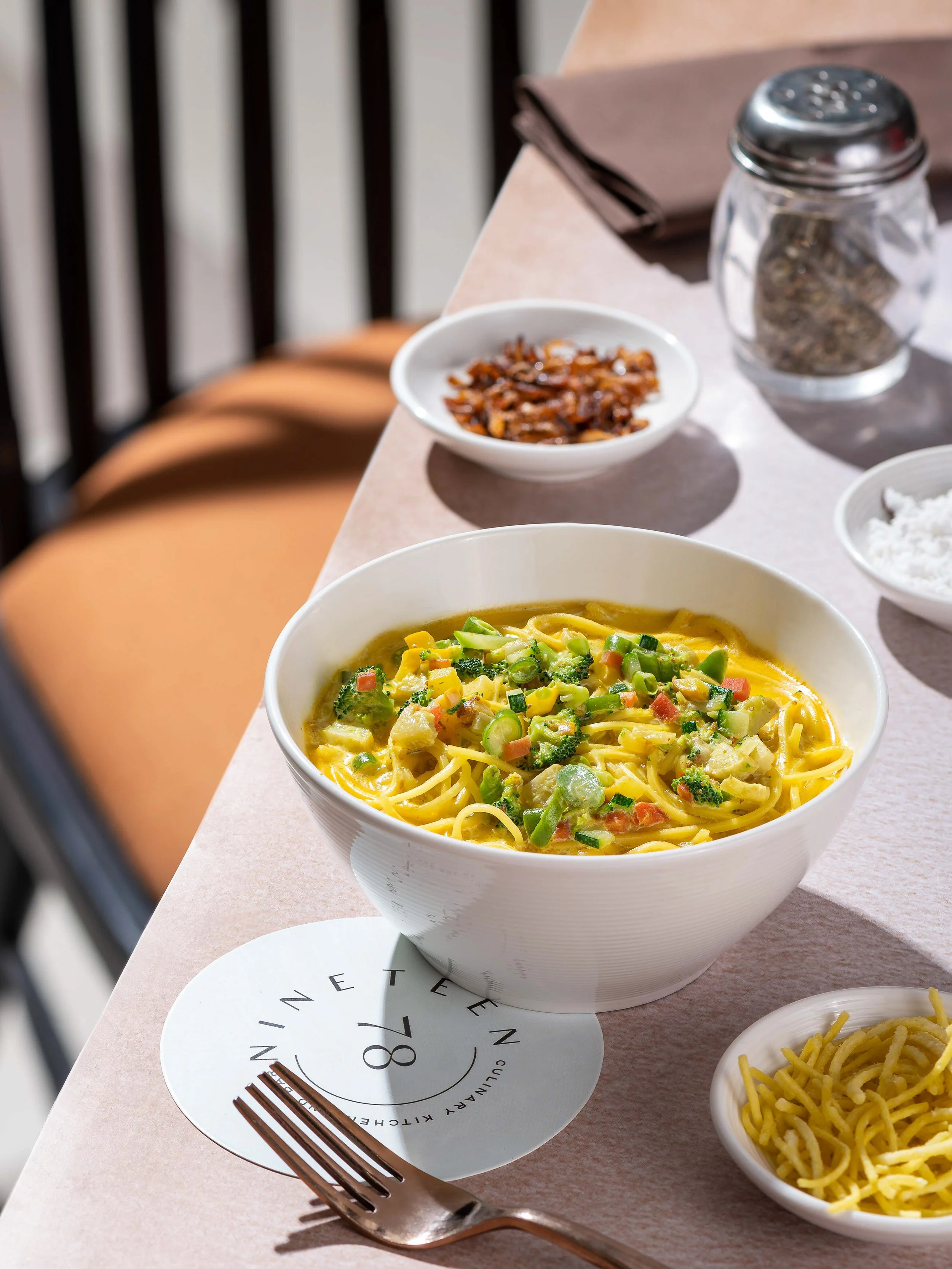 A bowl of noodles with broccoli and diced vegetables on a light pink table, with a small dish of crispy noodles, a glass jar of spices, a plate with a napkin, and a fork nearby.