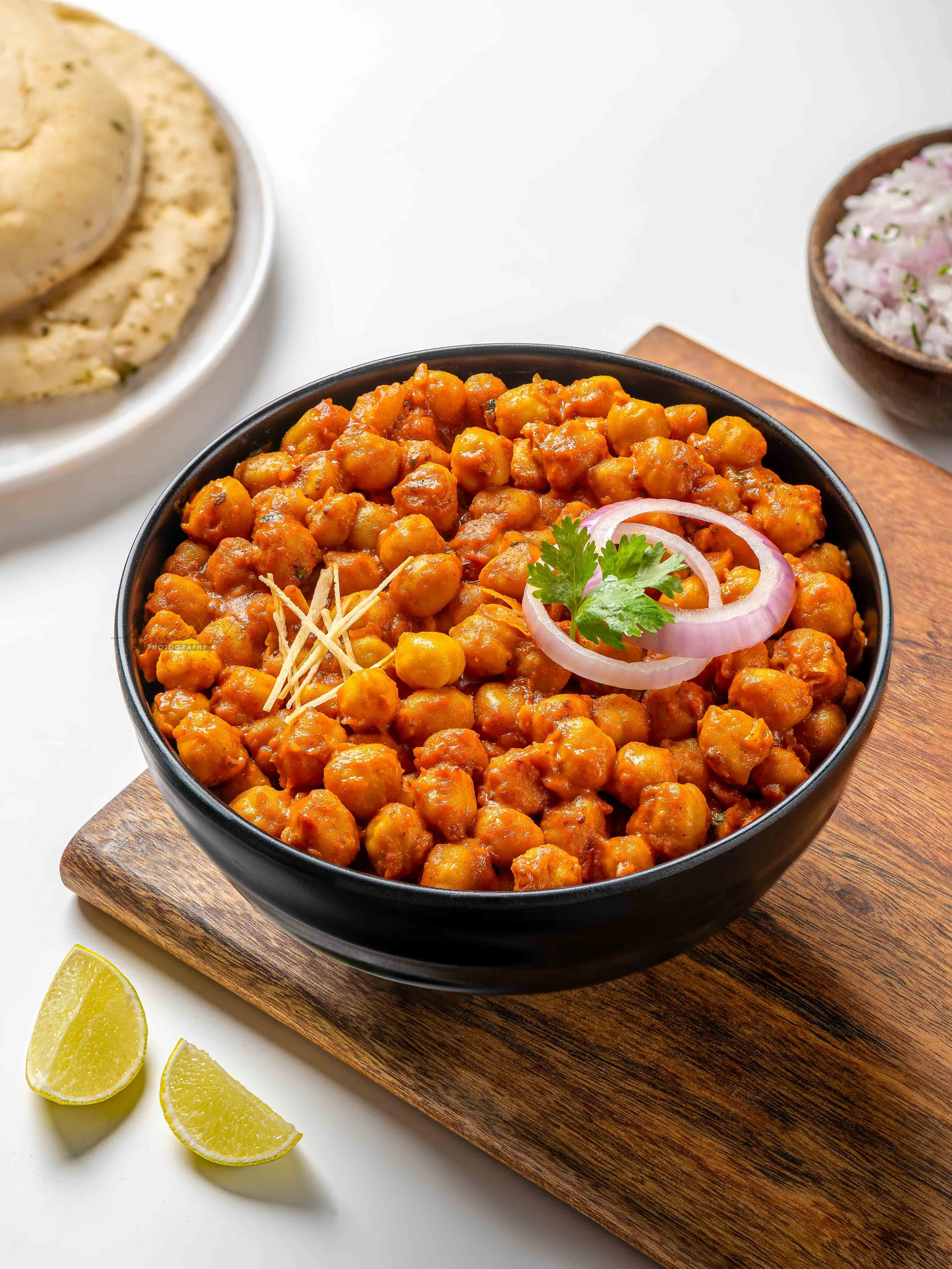 A bowl of spicy chickpea curry garnished with onion slices and cilantro, served on a wooden board with lemon wedges and naan bread in the background.