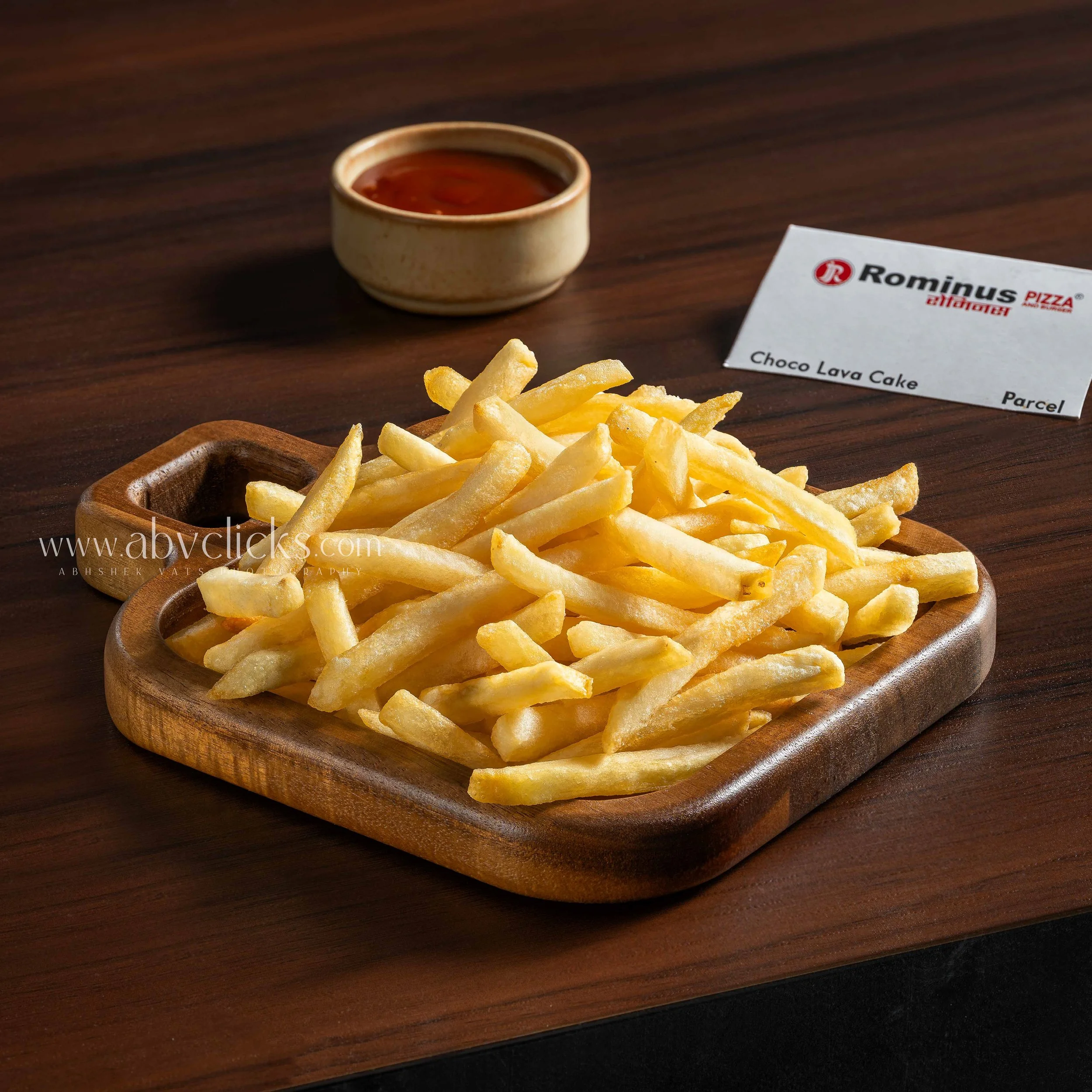 Small bowl of red sauce, plate of French fries, and a sign that reads 'Romnus Pizza, Choco Lava Cake, Parcel' on a wooden table.