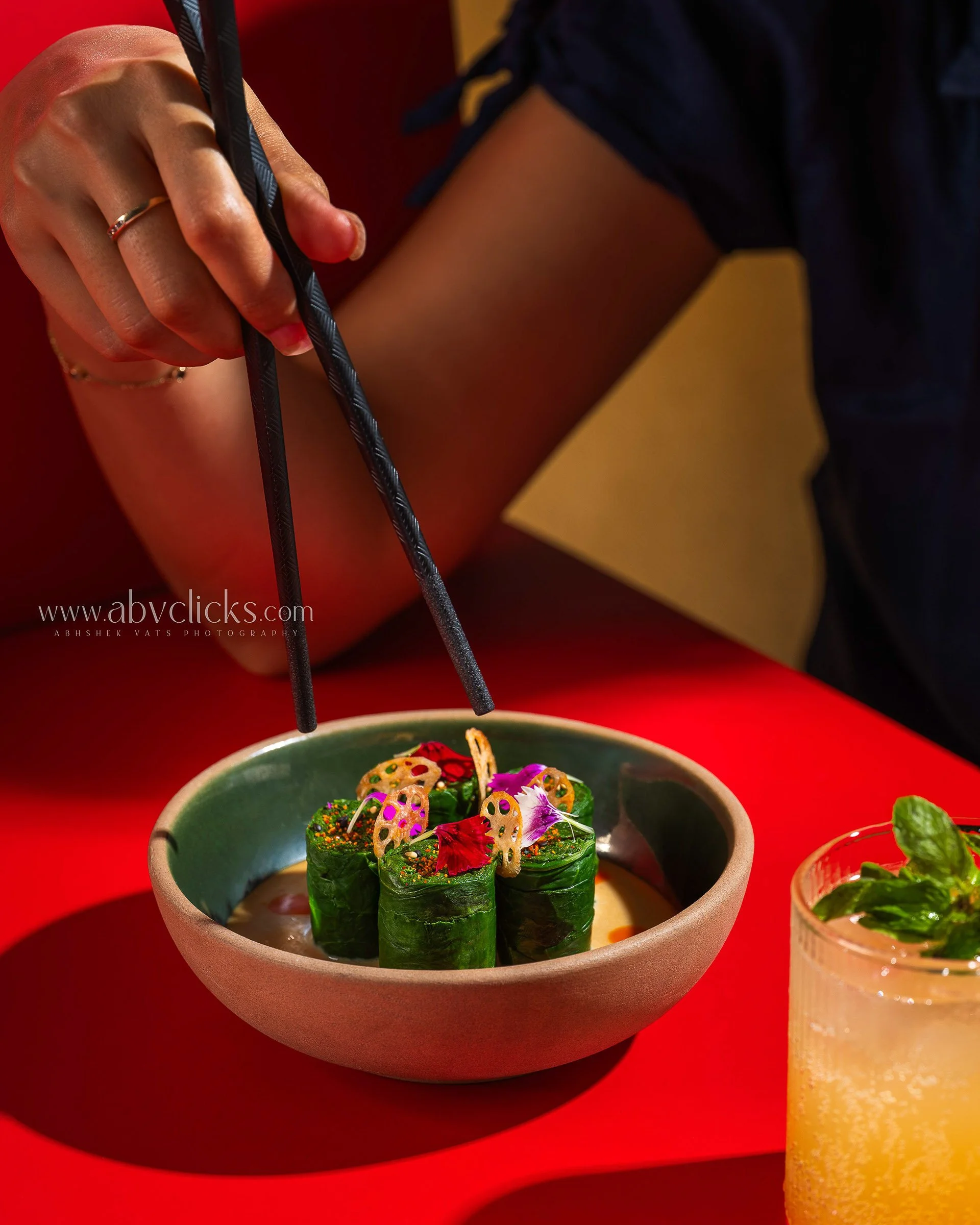 Close-up of cucumber rolls garnished with colorful edible flowers and lotus root chips in a green bowl on a red surface.
