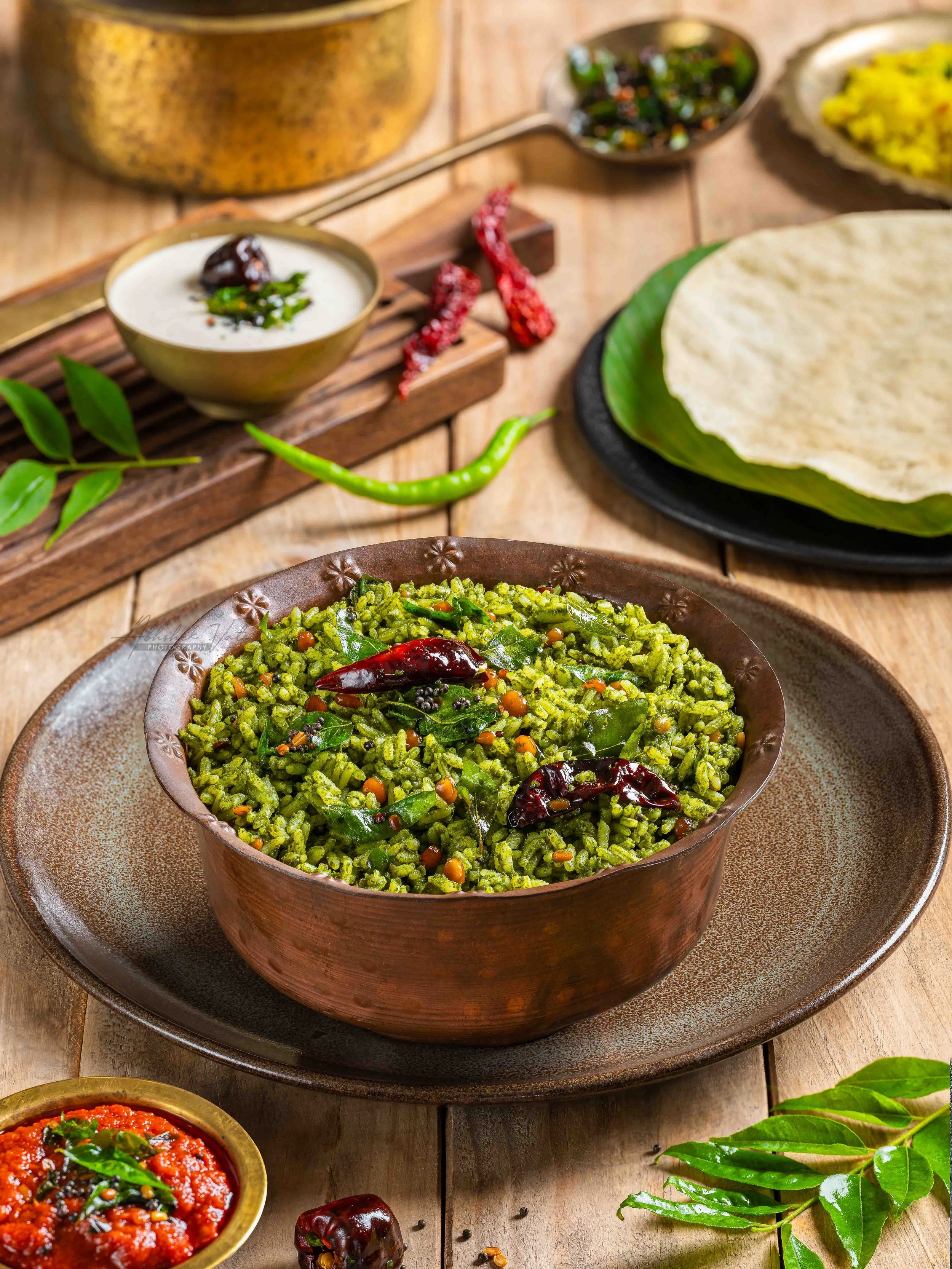 A bowl of cooked green rice with dried red chilies and spices, surrounded by small bowls of chutney, a leaf, and red chilies, on a wooden table.