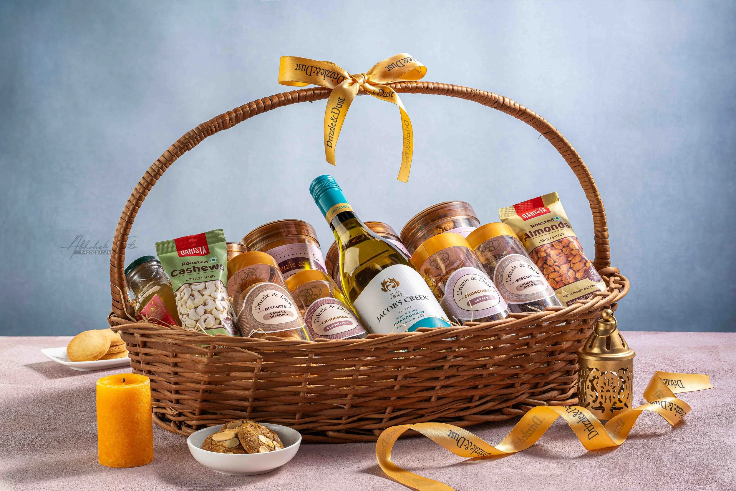 A wicker gift basket filled with assorted snacks such as roasted almonds, cashews, biscuits, and other treats, decorated with a yellow ribbon and set against a plain background.