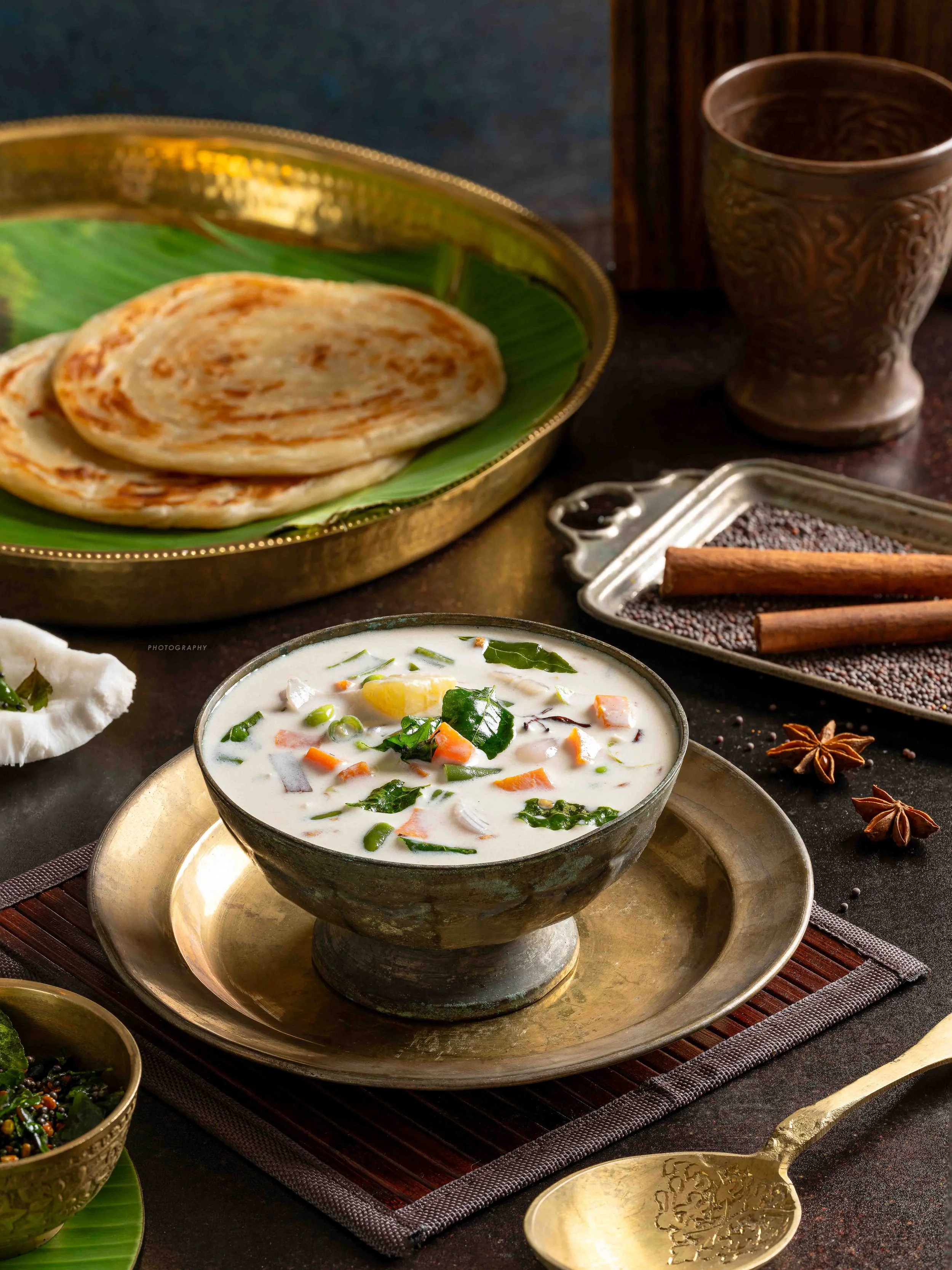 A bowl of Indian curry with vegetables, a plate of stuffed paratha on a green banana leaf, an ornate copper cup, a silver tray with black cumin seeds, and star anise on a dark table.