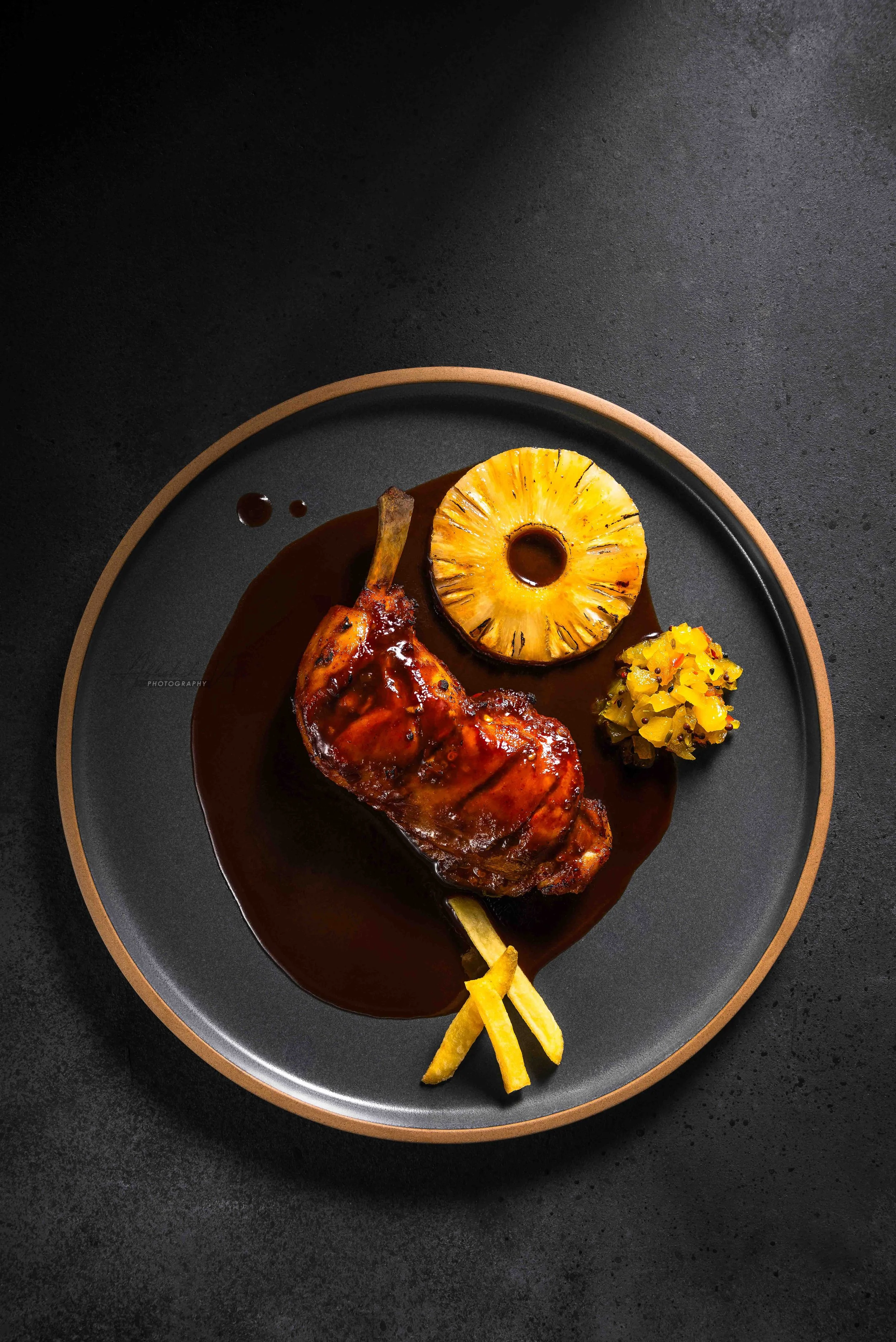 A plate with a piece of roasted meat topped with sauce, a grilled pineapple ring, a small portion of diced yellow vegetables, and two yellow fry-like garnishes on a black plate with a light brown border, placed on a dark textured surface.