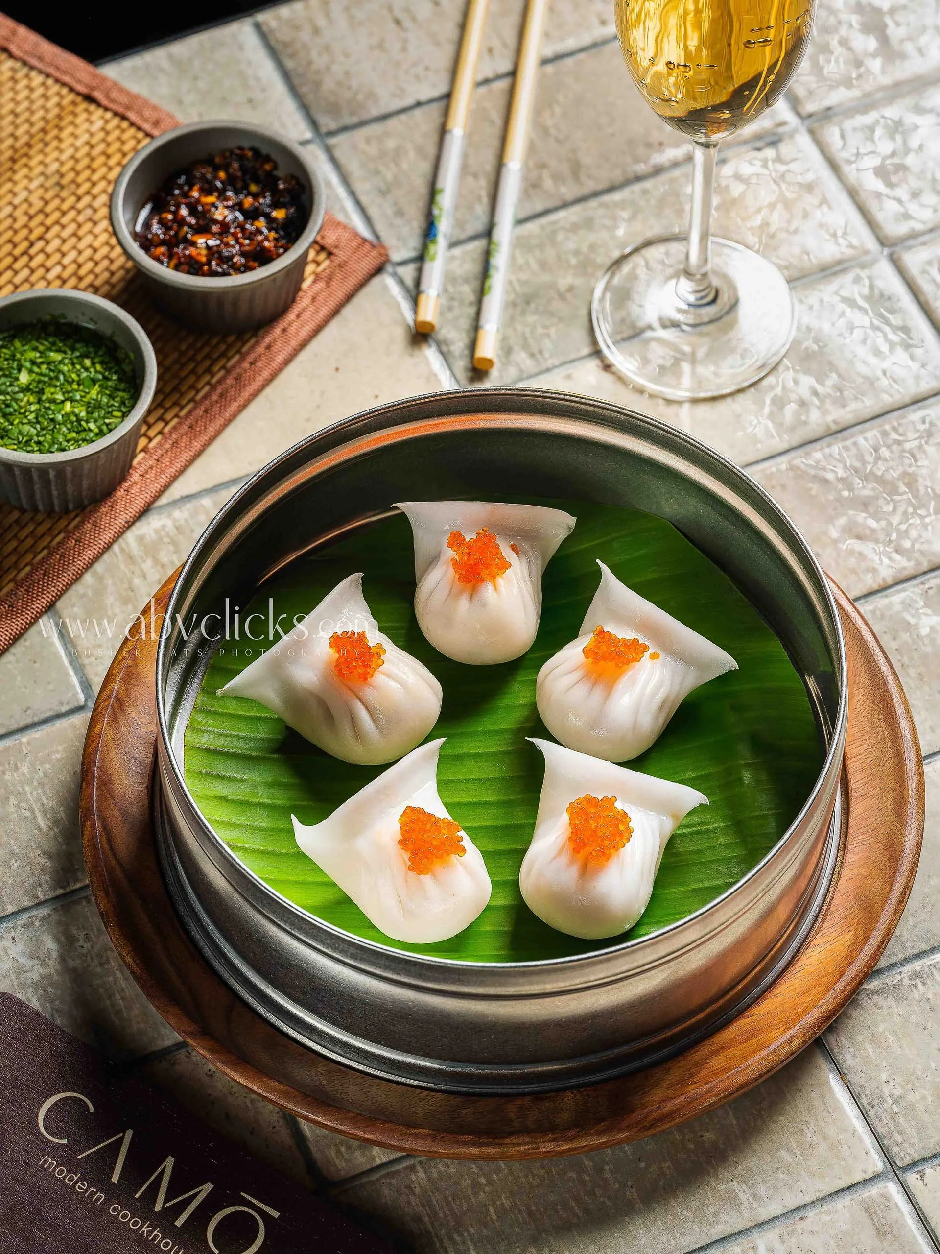 Steamed dim sum dumplings with orange fish roe on top in a traditional bamboo steamer lined with green leaves, placed on a wooden tray. Side dishes include chopped green and dark sauces in small bowls, chopsticks, a glass of white wine, and a tiled t