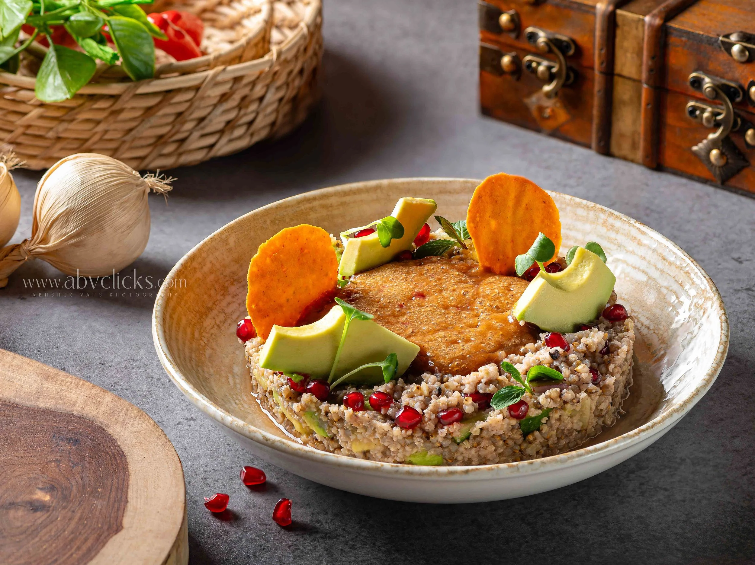 A bowl of vegetarian dish with rice, topped with avocado slices, pomegranate seeds, and crispy plantain chips, served on a gray textured surface. Basket of vegetables and a wooden chest in the background.