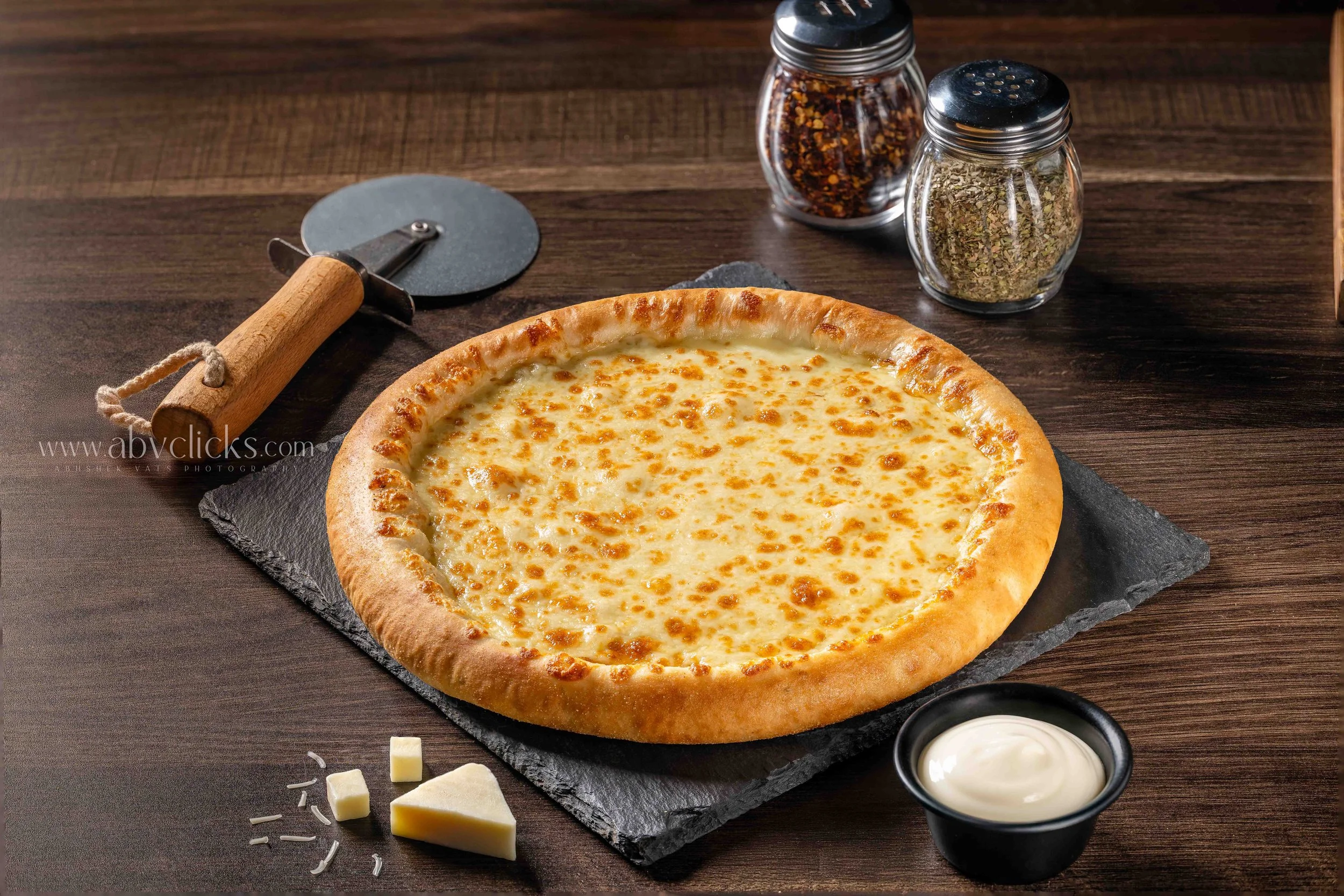 Cheese pizza on black slate with shredded cheese, black pepper, oregano, crushed red pepper, parmesan cheese, and a cup of ranch dressing on a dark wooden table.