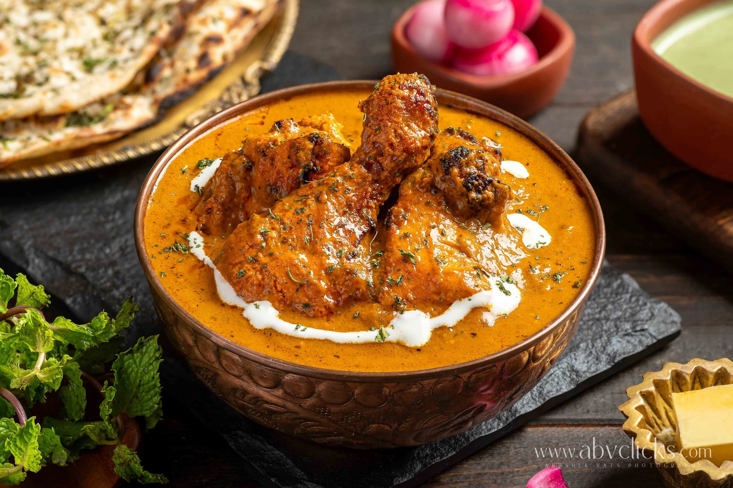 A bowl of Indian chicken curry with a creamy sauce, garnished with herbs, with naan bread, radishes, and a bowl of green sauce in the background.