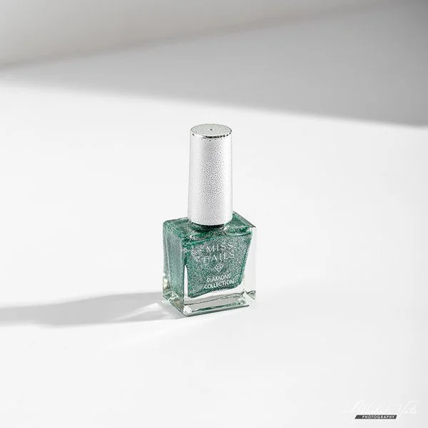 A square bottle of green nail polish with a silver cap, labeled 'Miss Nails Diamond Collection,' placed on a white surface with a gray background.