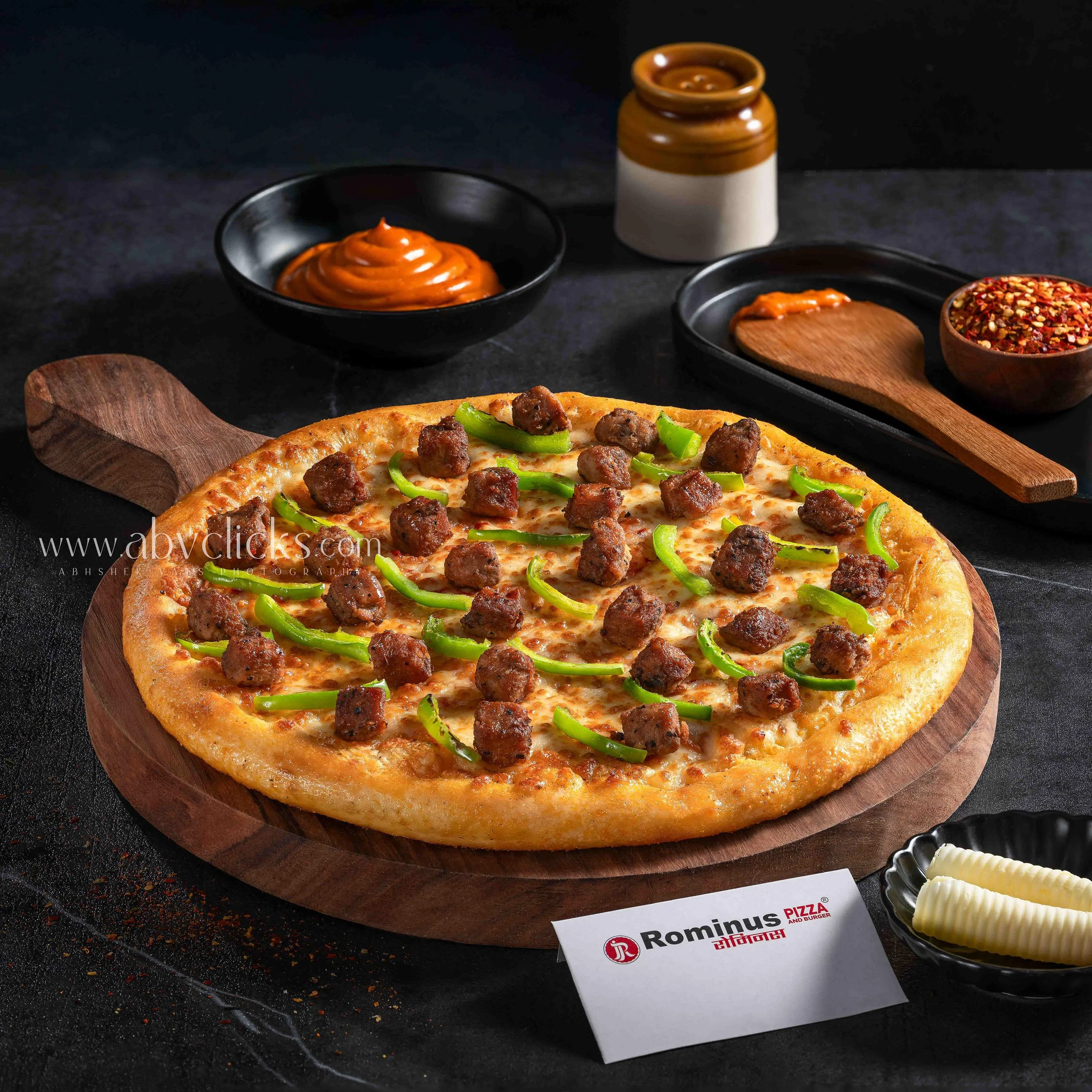 Close-up of a freshly baked pizza on a wooden pizza peel, topped with ground meat and green bell peppers, with a side of spicy red pepper flakes and a small container of butter, set on a black surface with scattered spices and a white paper with red 