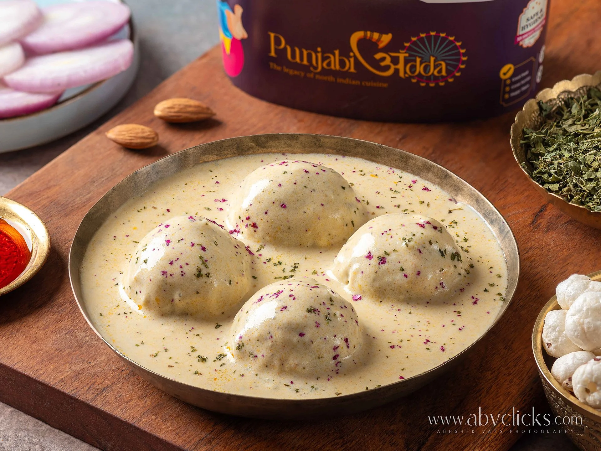 A traditional Indian dish called Ras Malai served in a metal bowl, garnished with dried rose petals and sprinkled with cardamom powder, with ingredients like almonds and dried herbs nearby on a wooden surface.