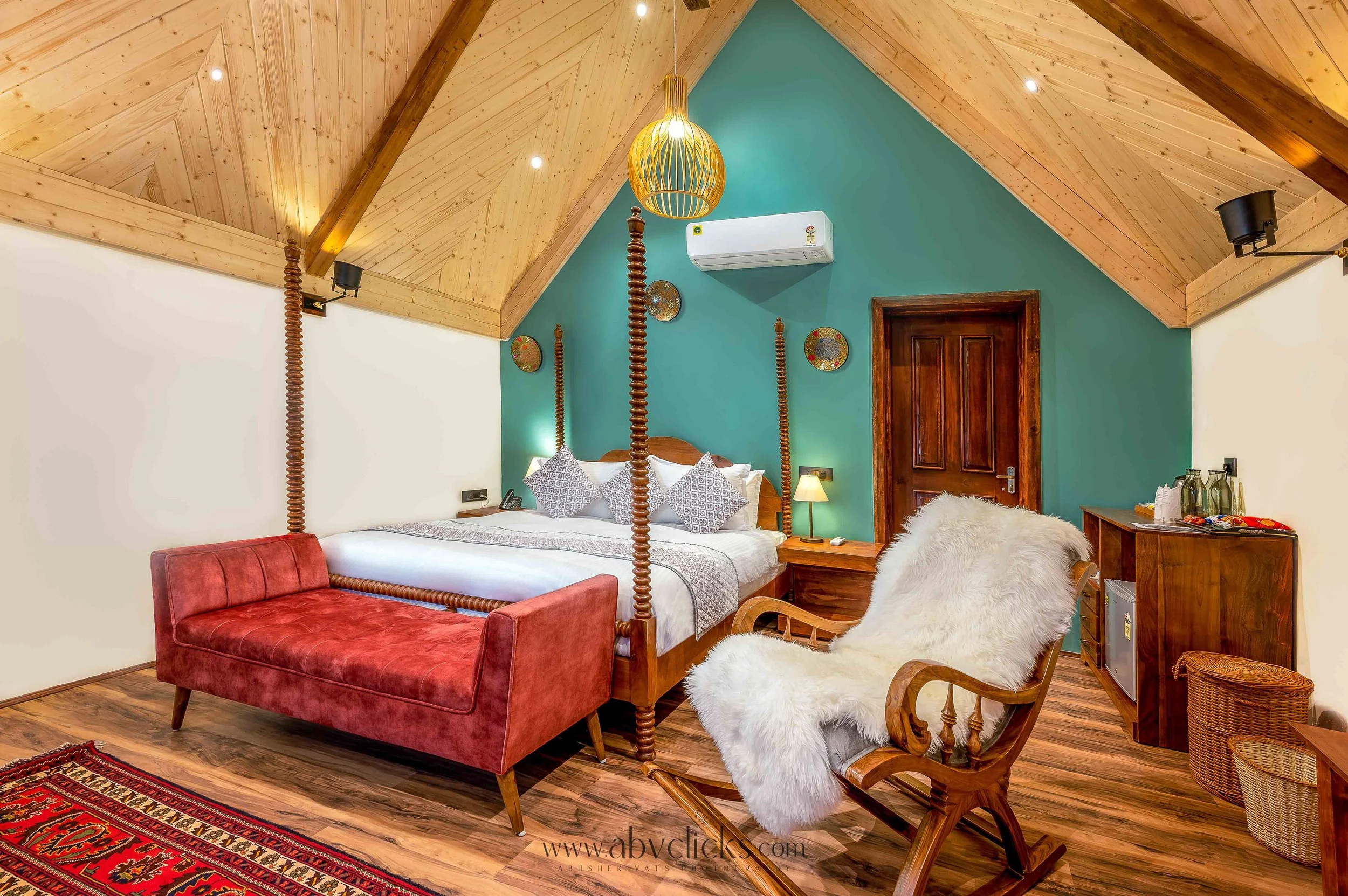 Interior of a bedroom with a wooden vaulted ceiling, turquoise accent wall, four-poster bed, red velvet bench, white furry armchair, wooden dining chairs, and various decorative plates on the wall.