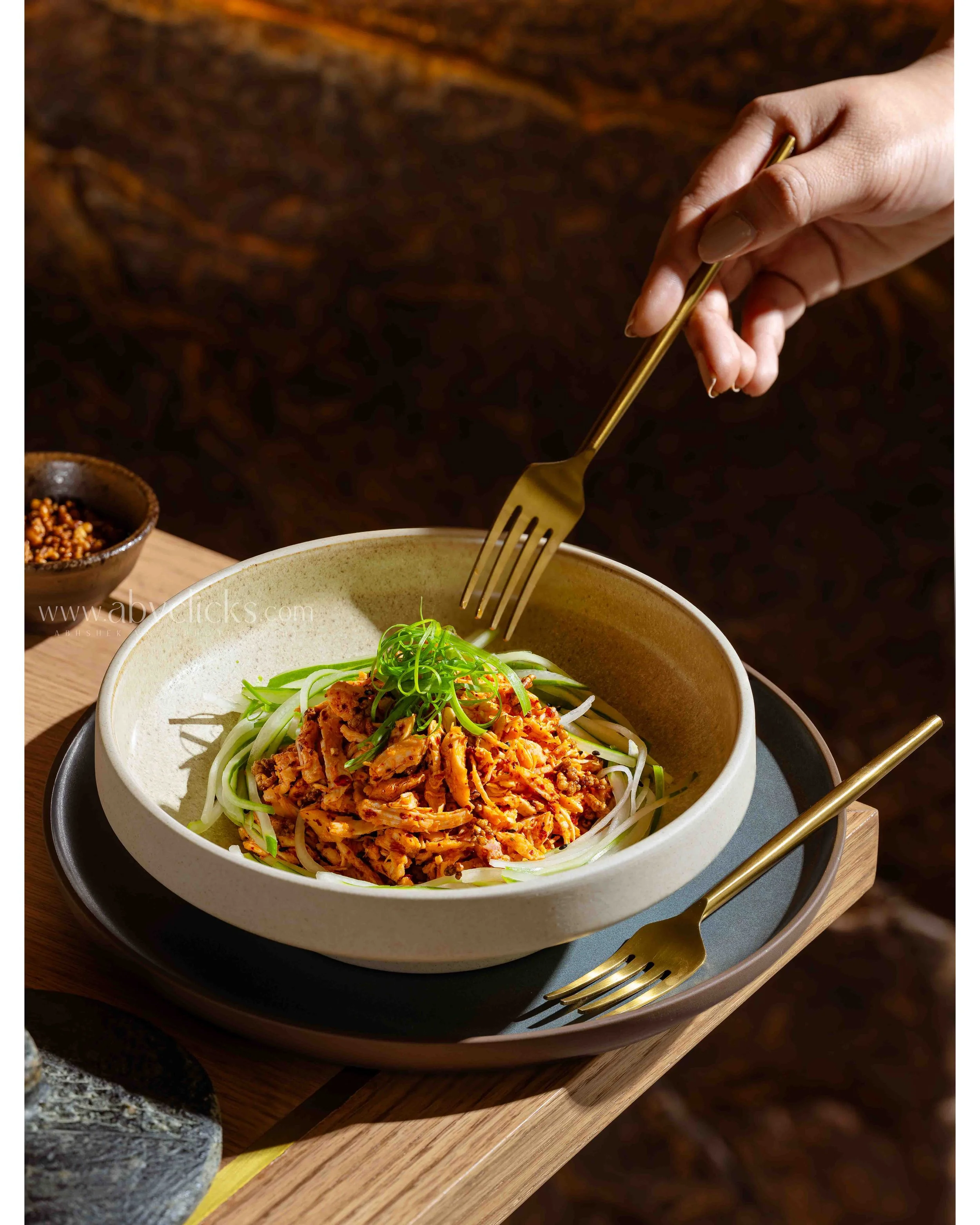 A bowl of Asian-inspired noodle dish topped with shredded meat and green garnishes. The bowl is placed on a black tray on a wooden table, with a gold fork and small bowl of peppercorns nearby in a cozy, dimly lit setting.