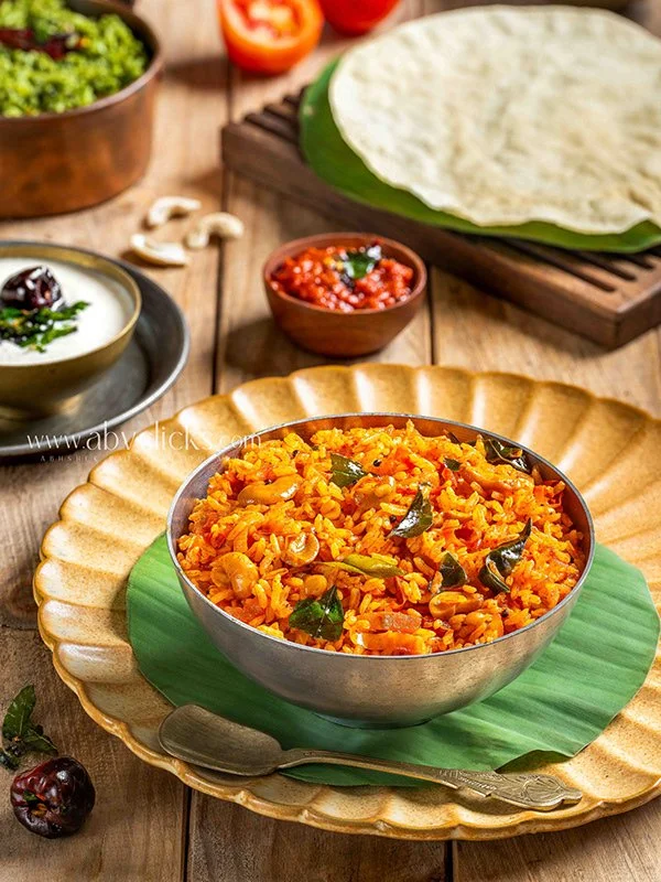 Bowl of spicy vegetable and rice pulao garnished with curry leaves, served on a decorative plate with green leaf underlay, surrounded by small bowls of tomato salsa, raita, and additional ingredients on a rustic wooden table.