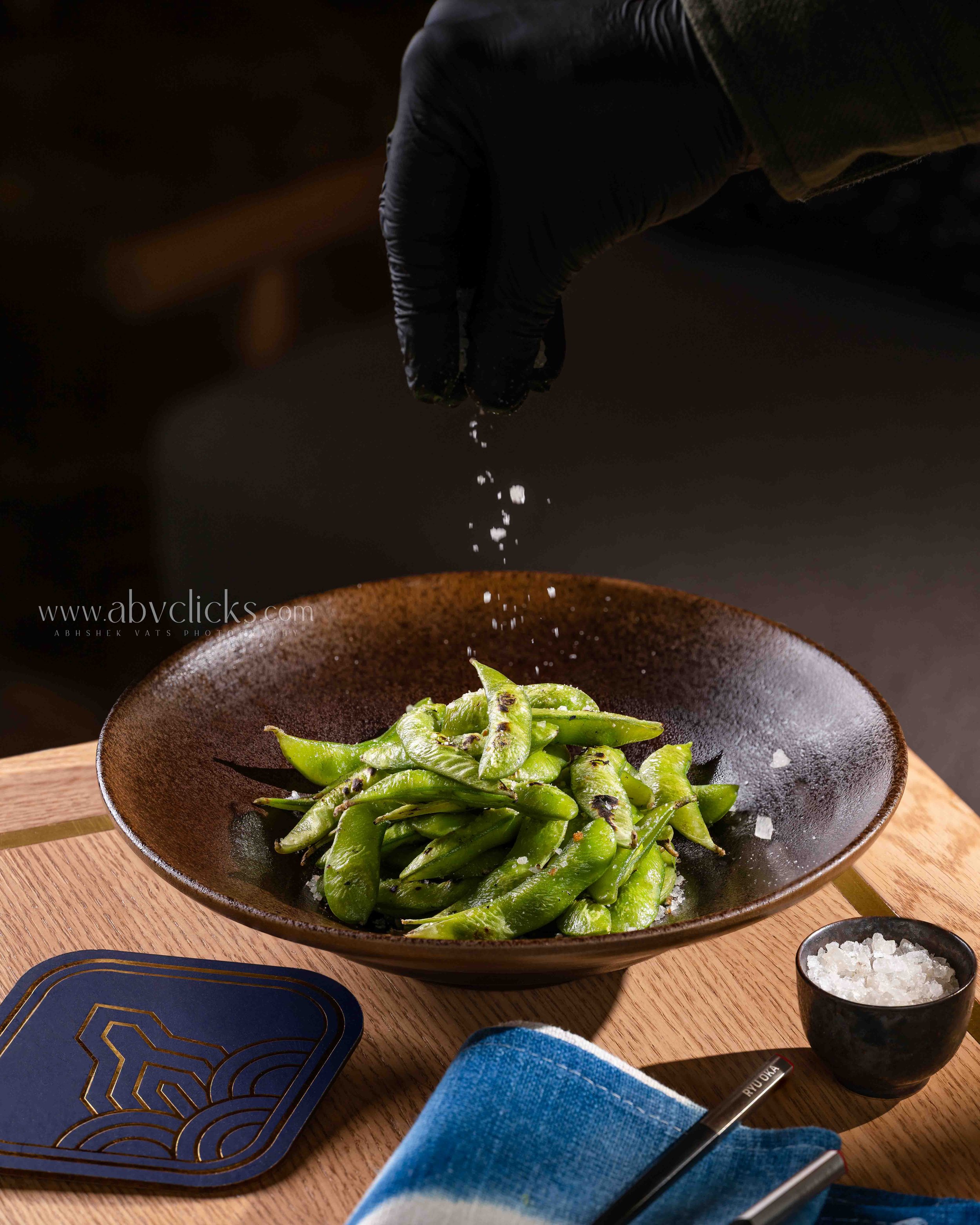 A person wearing a black glove sprinkling salt over grilled green peppers in a dark brown ceramic bowl on a wooden table, with other bowls and utensils nearby.