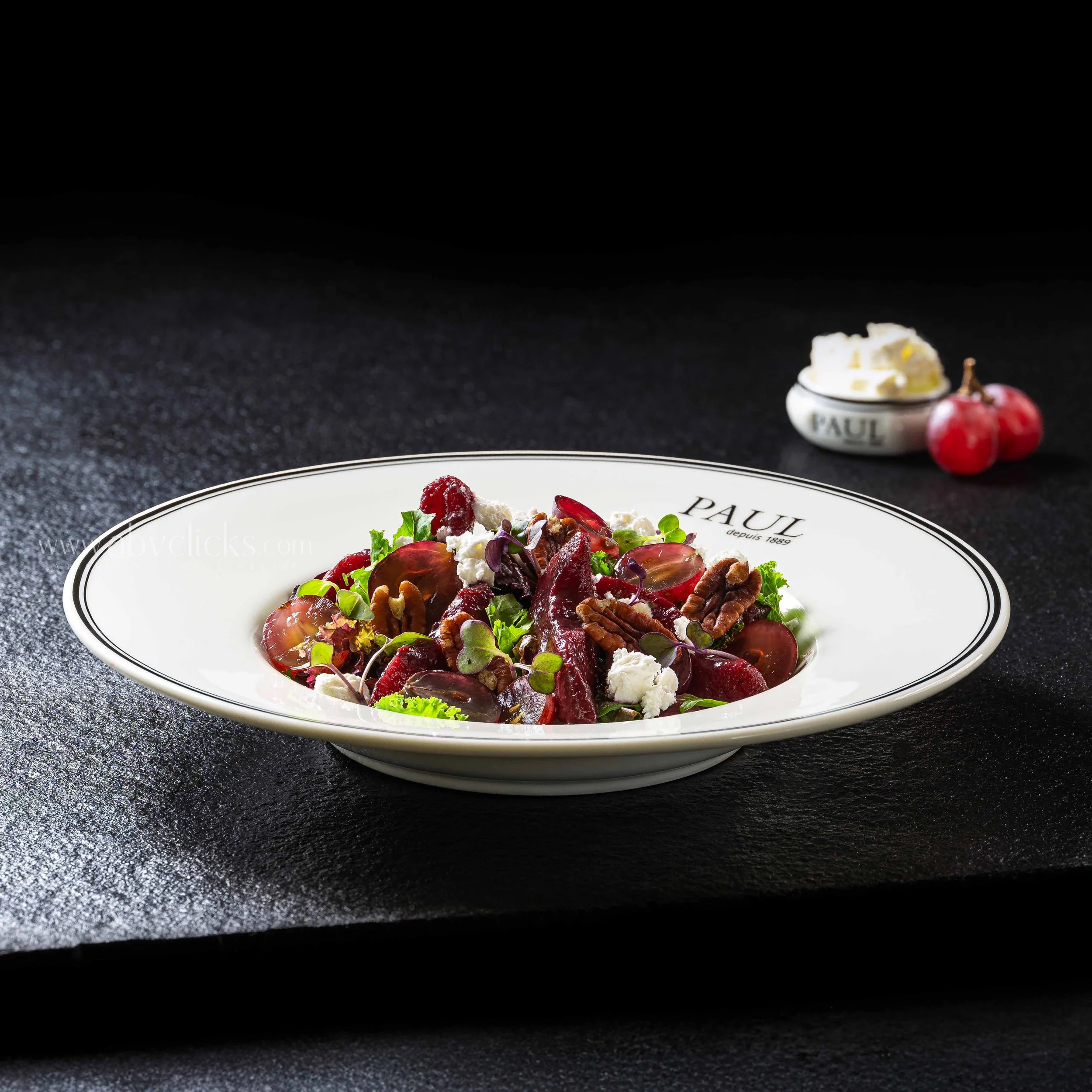 A bowl of salad with mixed greens, sliced beets, cheese crumbles, and nuts on a black textured surface, with a small dish of butter and a few cherries in the background.