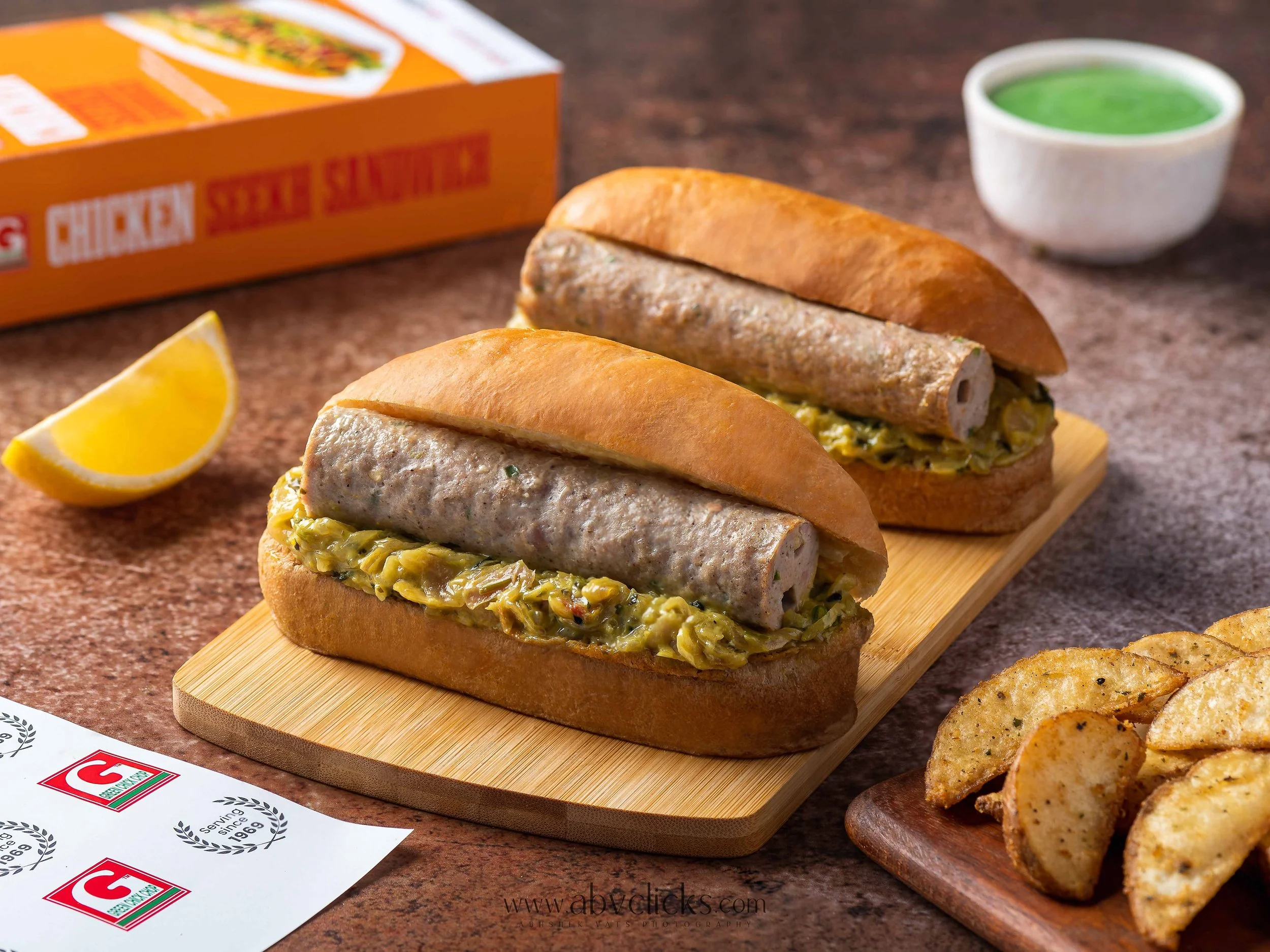 A hot dog with a sausage, relish, and bun on a wooden cutting board, lemon wedge, potato chips, a cup of green sauce, and a box of frozen potato products on a countertop.