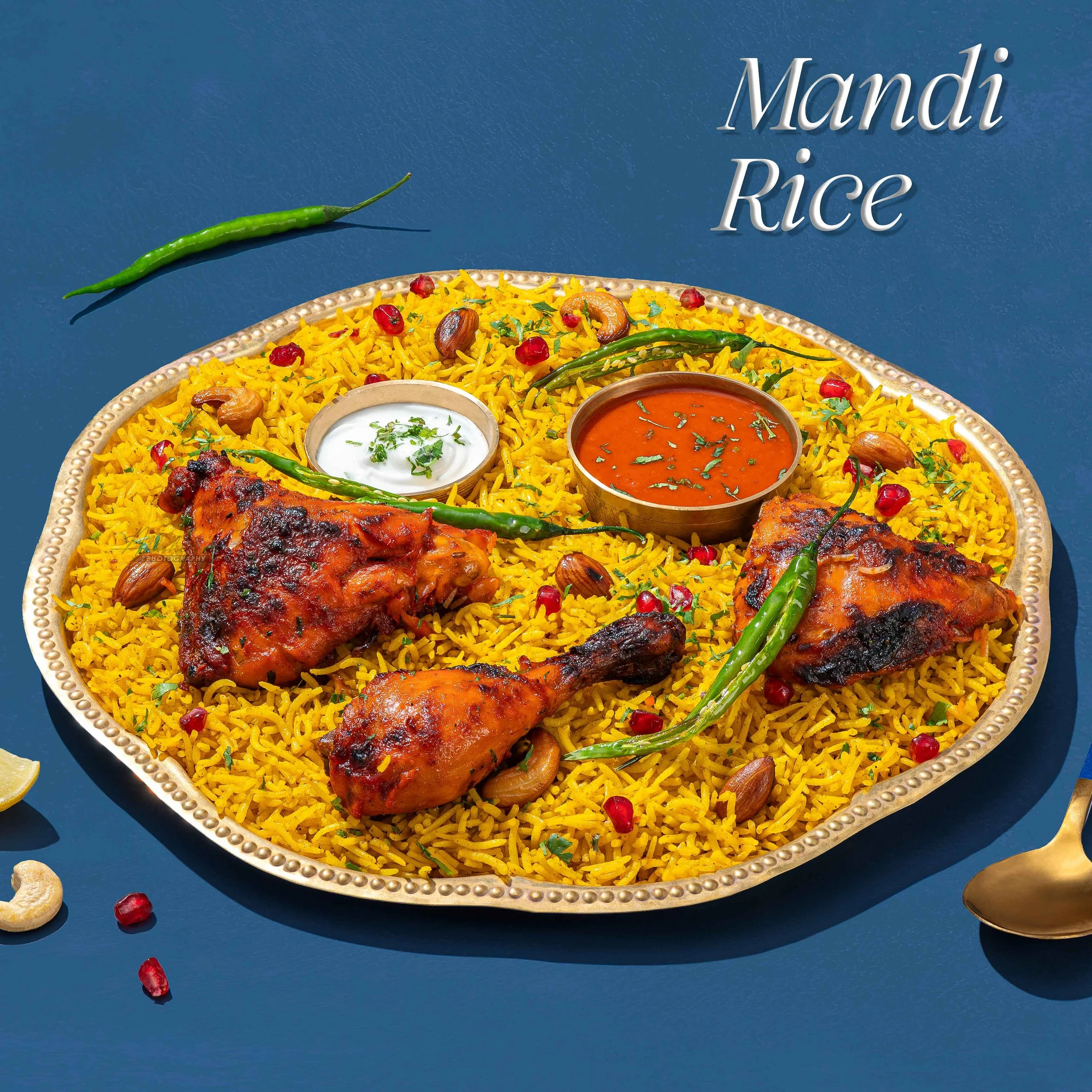 Close-up of a tray of traditional Indian yellow rice garnished with pomegranate seeds, cashews, and cilantro, with two bowls of curry and a side of green chili peppers, on a blue background. The text 'Mandi Rice' is displayed in the background.