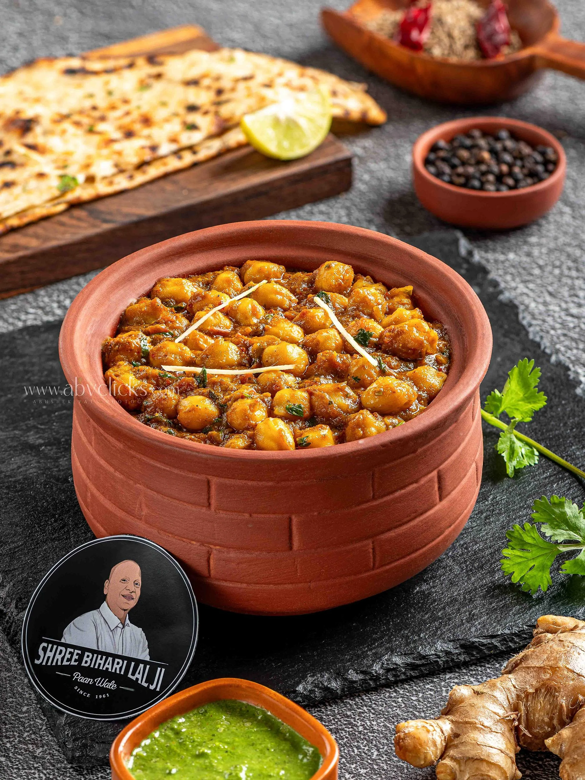 Close-up of a terracotta bowl filled with chana masala, surrounded by small bowls of green chutney, black pepper, and a plate of flatbread garnished with lemon wedge and herbs, set on a dark textured surface.