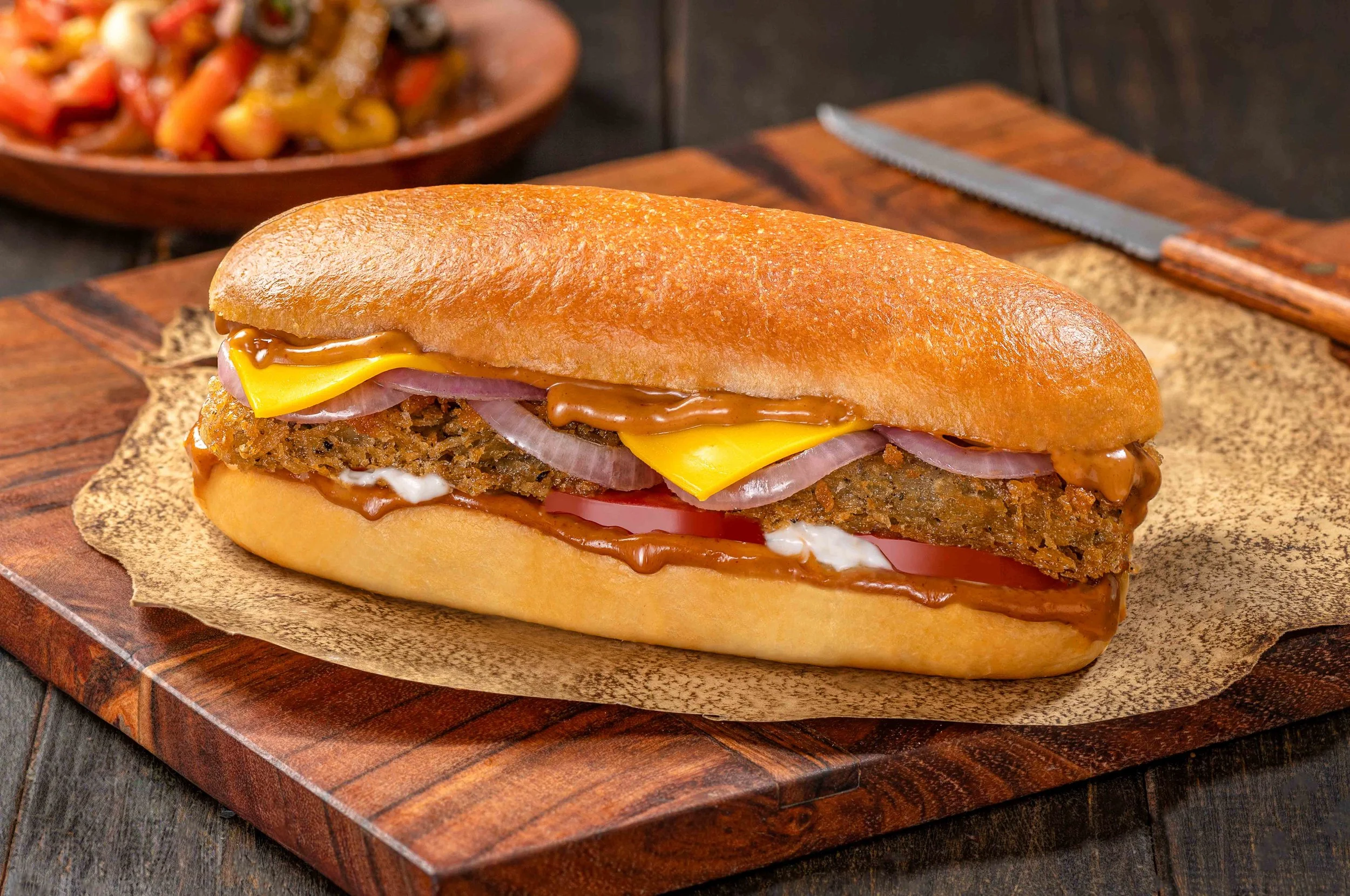 A close-up of a sandwich with fried chicken filet, cheddar cheese, red onion, tomato, and barbecue sauce in a baguette-style bun on a wooden serving board.