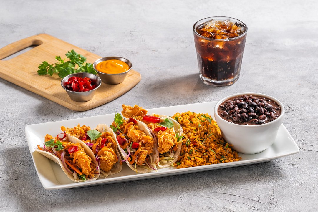 Three fish tacos with fried fish, cabbage slaw, and red peppers on a rectangular white plate, served with a side of Mexican rice and black beans, and two small bowls of sauces, with a glass of cola and ice, on a gray surface.